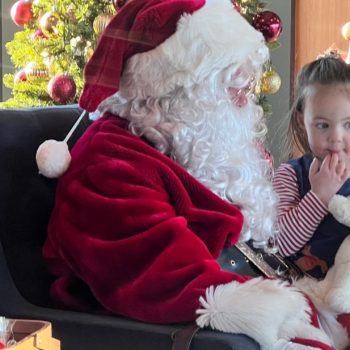 Young girl holding a stuffed animal sits quietly beside Santa Claus in a cozy holiday setting with a decorated Christmas tree in the background.