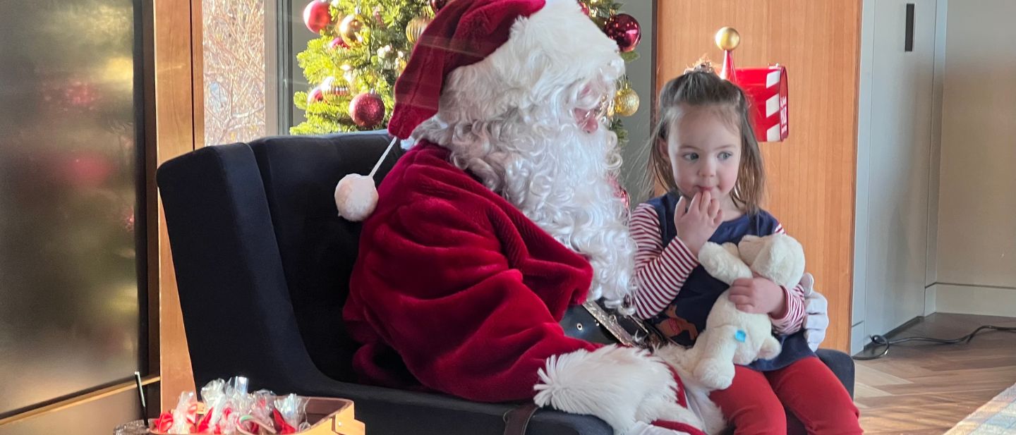 Young girl holding a stuffed animal sits quietly beside Santa Claus in a cozy holiday setting with a decorated Christmas tree in the background.
