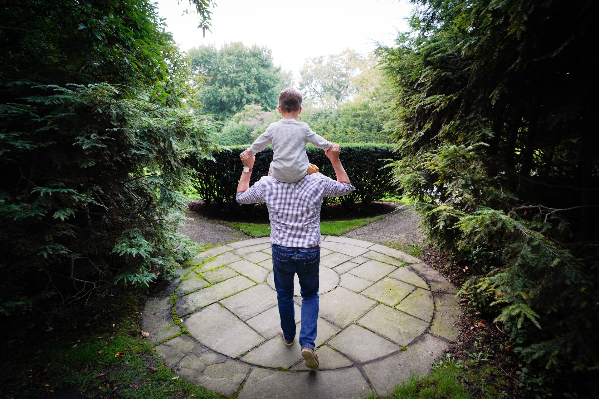 A father carries his child on his shoulders while walking through a circular stone path surrounded by hedges on the grounds at Ford House.