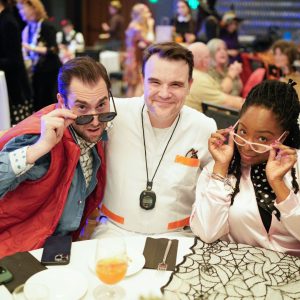 Three adults sit at a table. Two men are wearing costumes as Marty McFly and Doc Brown from Back to the Future and one woman is wearing a Pink Lady costume.