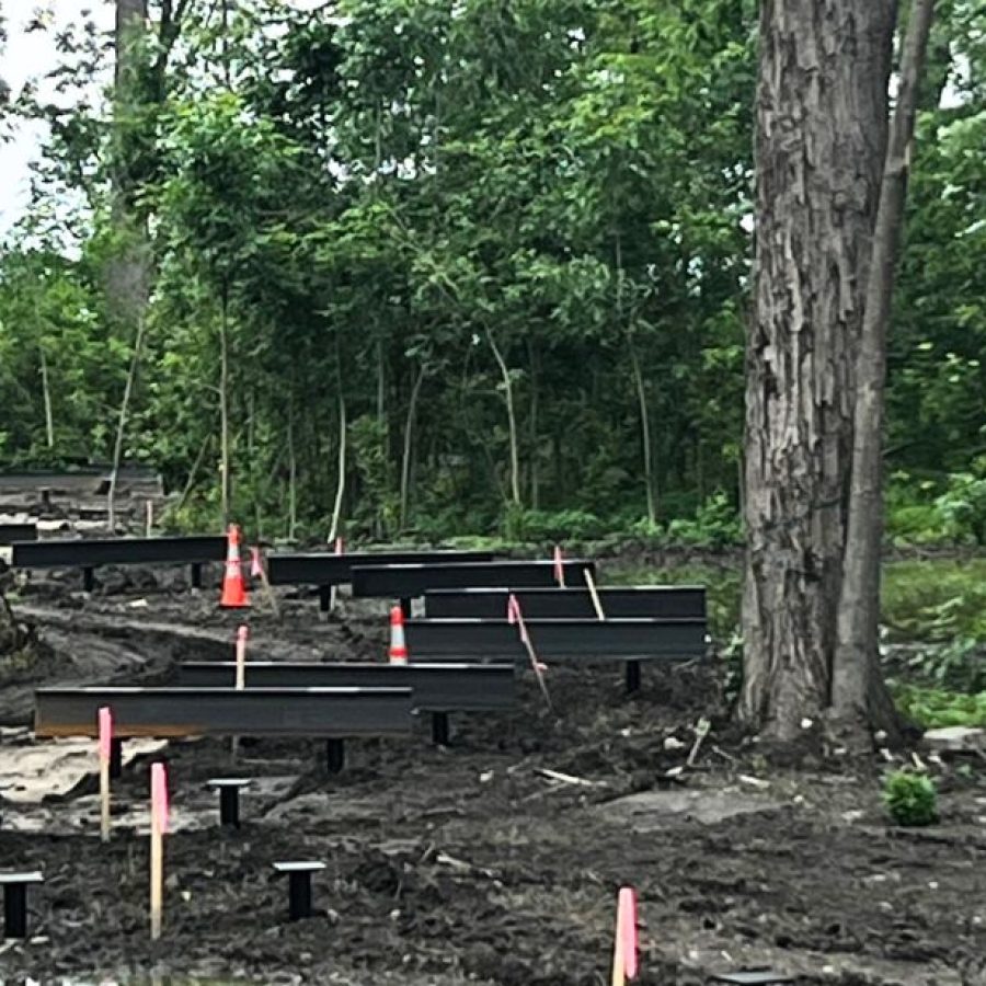 Construction progress on an elevated boardwalk through the wooded wetland near Ford House's Visitor Center, with black support beams and pink construction markers rising from muddy ground and reflective water, surrounded by tall trees and lush greenery.