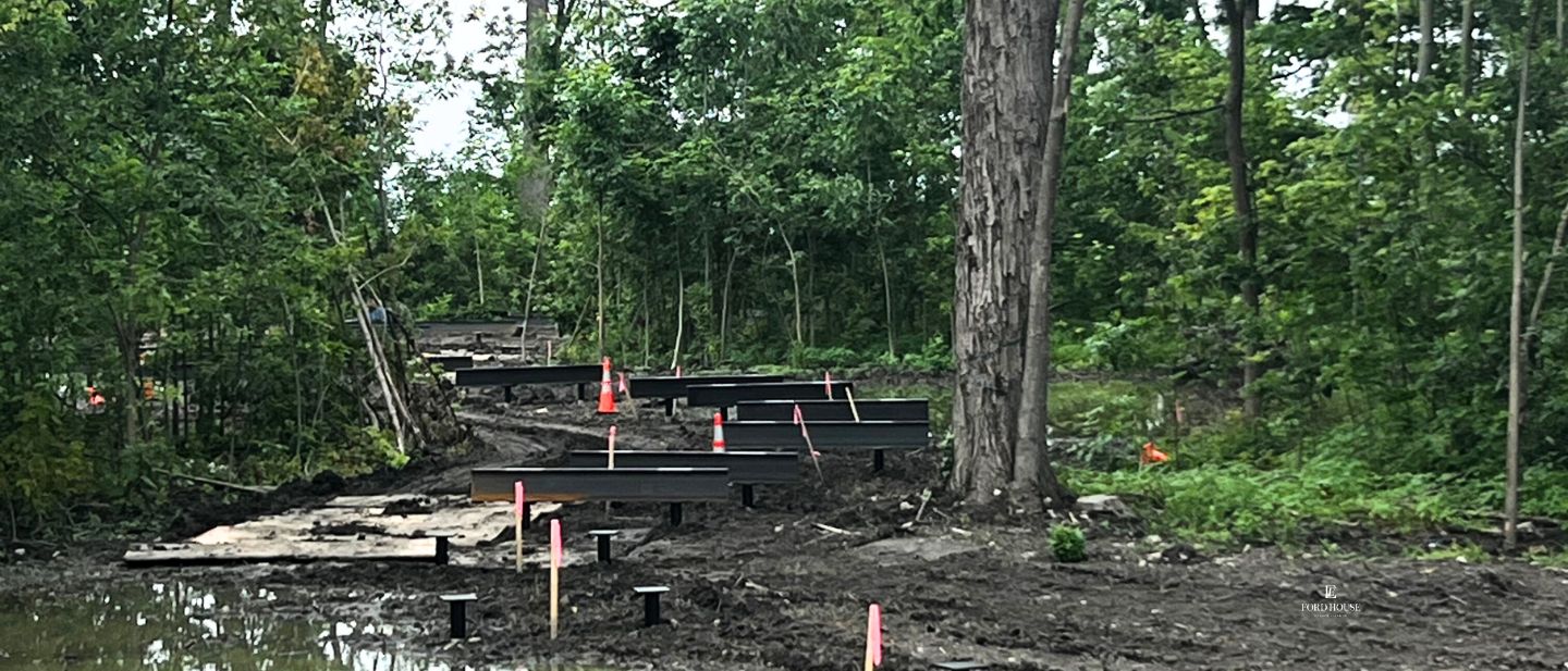 Construction progress on an elevated boardwalk through the wooded wetland near Ford House's Visitor Center, with black support beams and pink construction markers rising from muddy ground and reflective water, surrounded by tall trees and lush greenery.