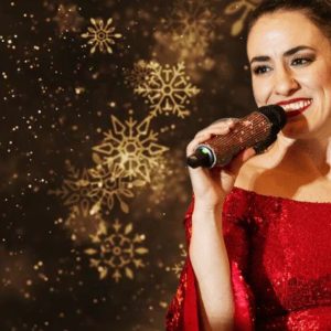 Vocalist Vanessa Carr smiles while performing in a red sequined gown against a warm background of glowing golden snowflake lights at Ford House’s Christmas Concert Over the Cove.