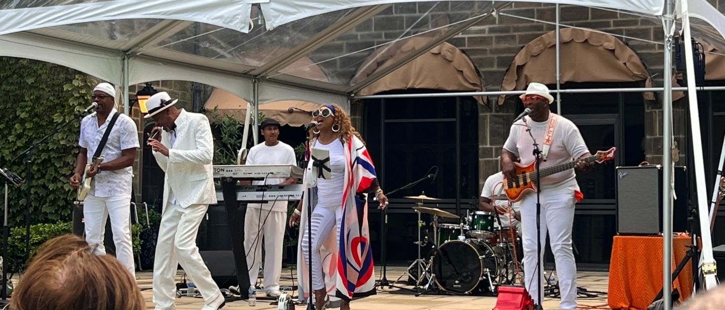 The Dave Hamilton Band performs live outdoors at Ford House, with vocalists, guitars, drums, and keyboard under a clear canopy on a sunny day.