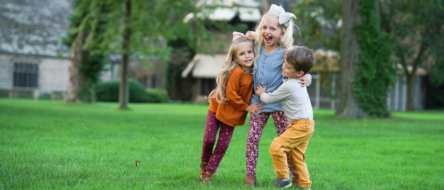 Three young children laughing and hugging while playing on the lush green lawn at Ford House, with the historic estate in the background.