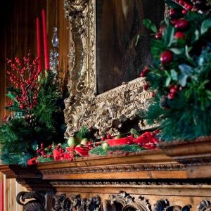 Holiday greenery, red berries, and candles decorate a carved wood mantel with an ornate frame inside the Main Residence at Ford House, part of the Main Residence Holiday Guided Tour.