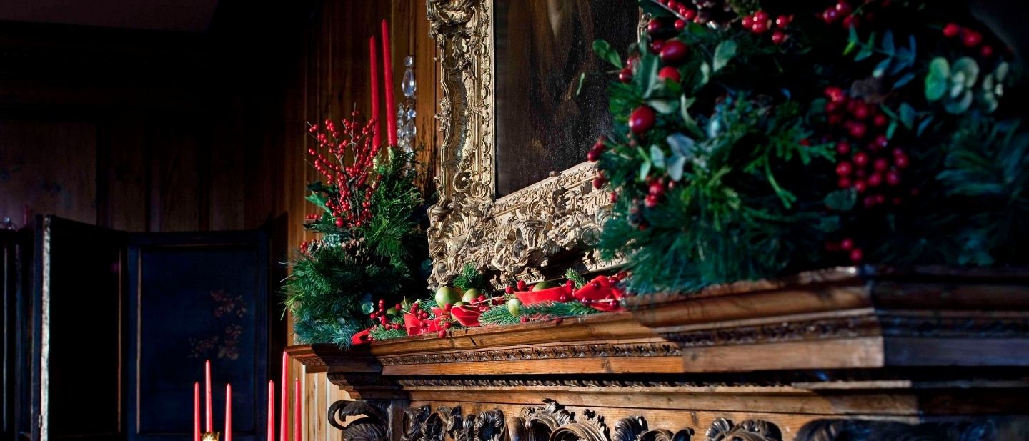 Holiday greenery, red berries, and candles decorate a carved wood mantel with an ornate frame inside the Main Residence at Ford House, part of the Main Residence Holiday Guided Tour.