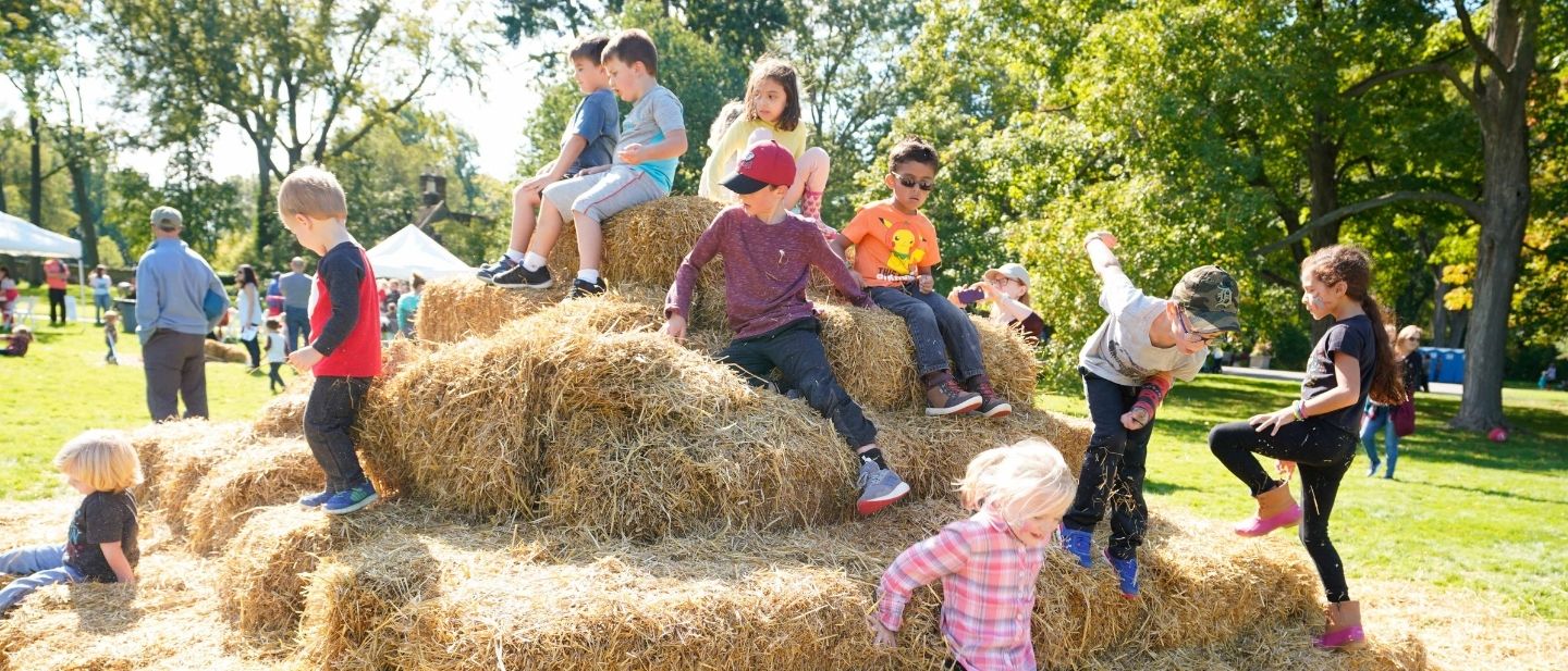 Children play on a large haystack during Harvest Day at Ford House, surrounded by fall activities and families enjoying the sunny outdoor festival.