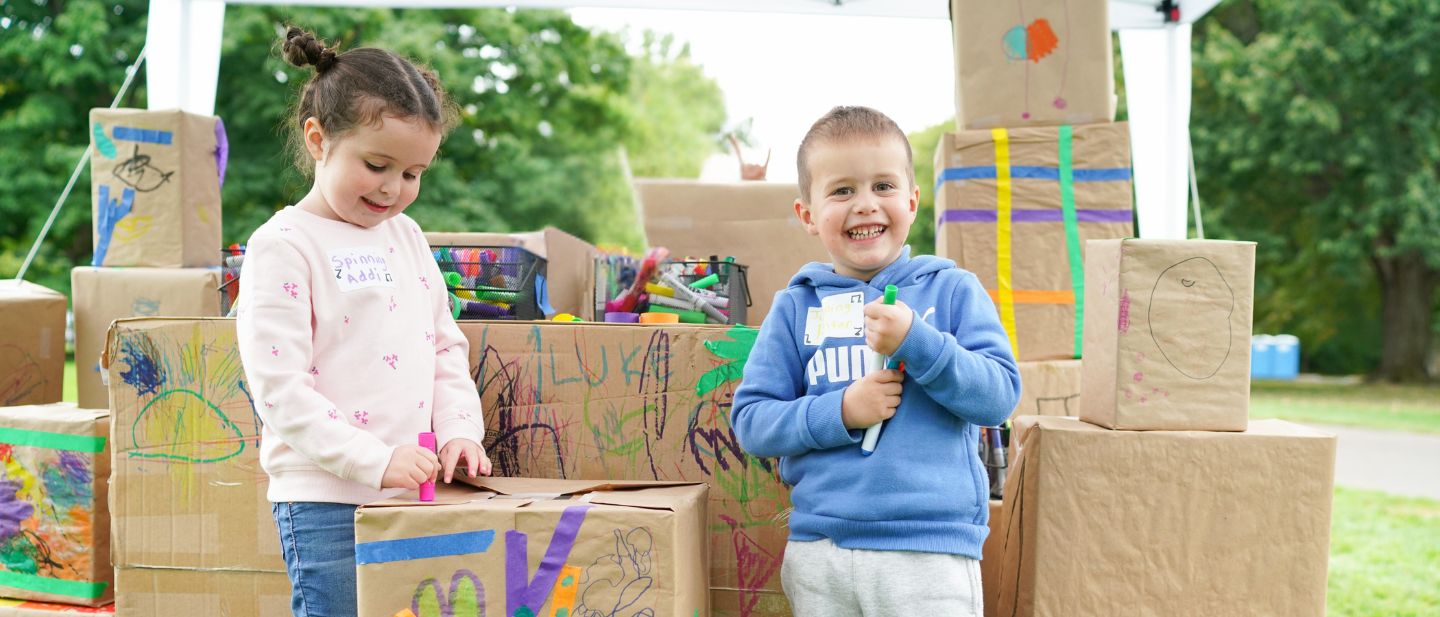 A young girl and boy decorating cardboard boxes with markers at Ford House’s Story Festival, surrounded by colorful art supplies outdoors.