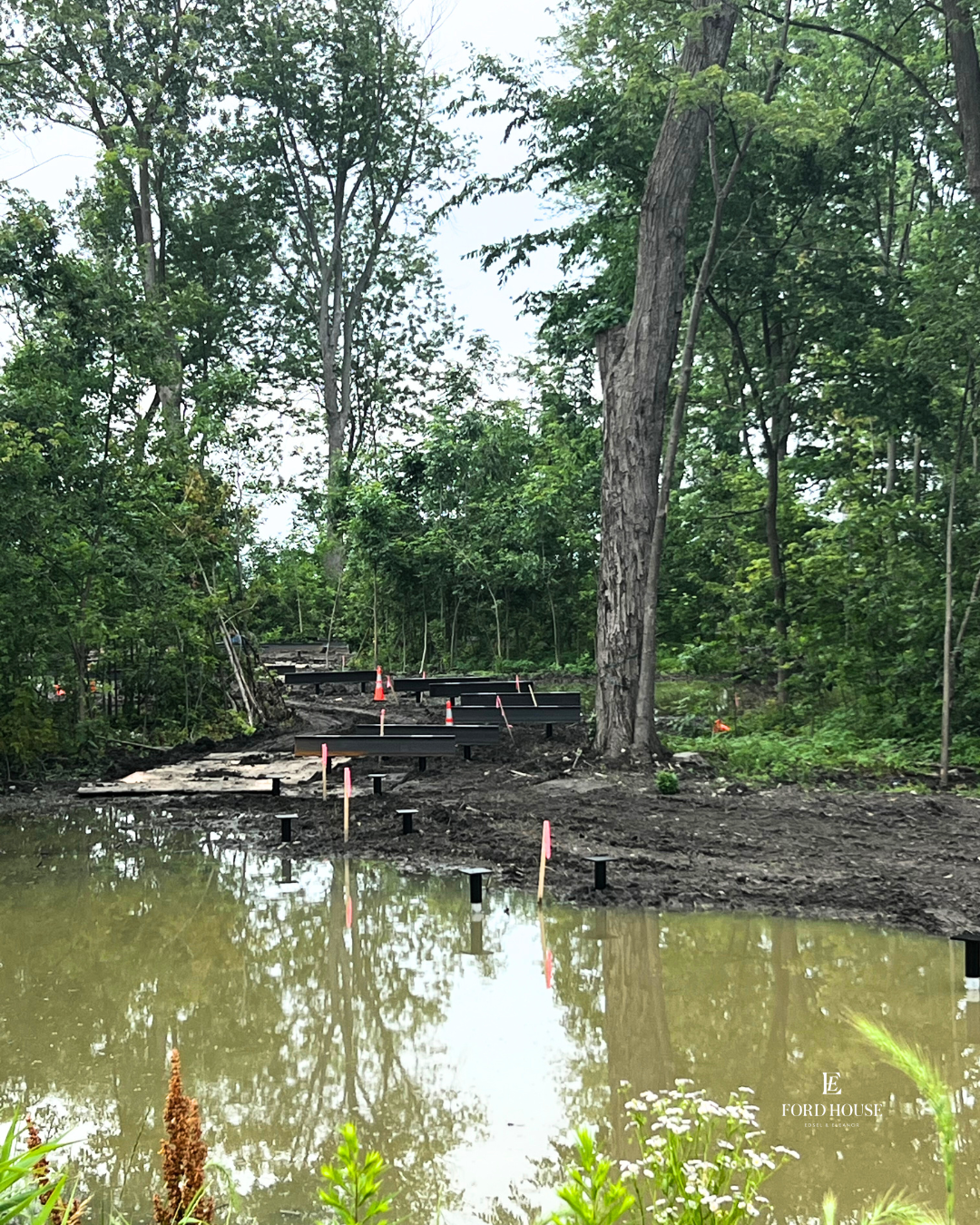 Construction progress on an elevated boardwalk through the wooded wetland near Ford House's Visitor Center, with black support beams and pink construction markers rising from muddy ground and reflective water, surrounded by tall trees and lush greenery.