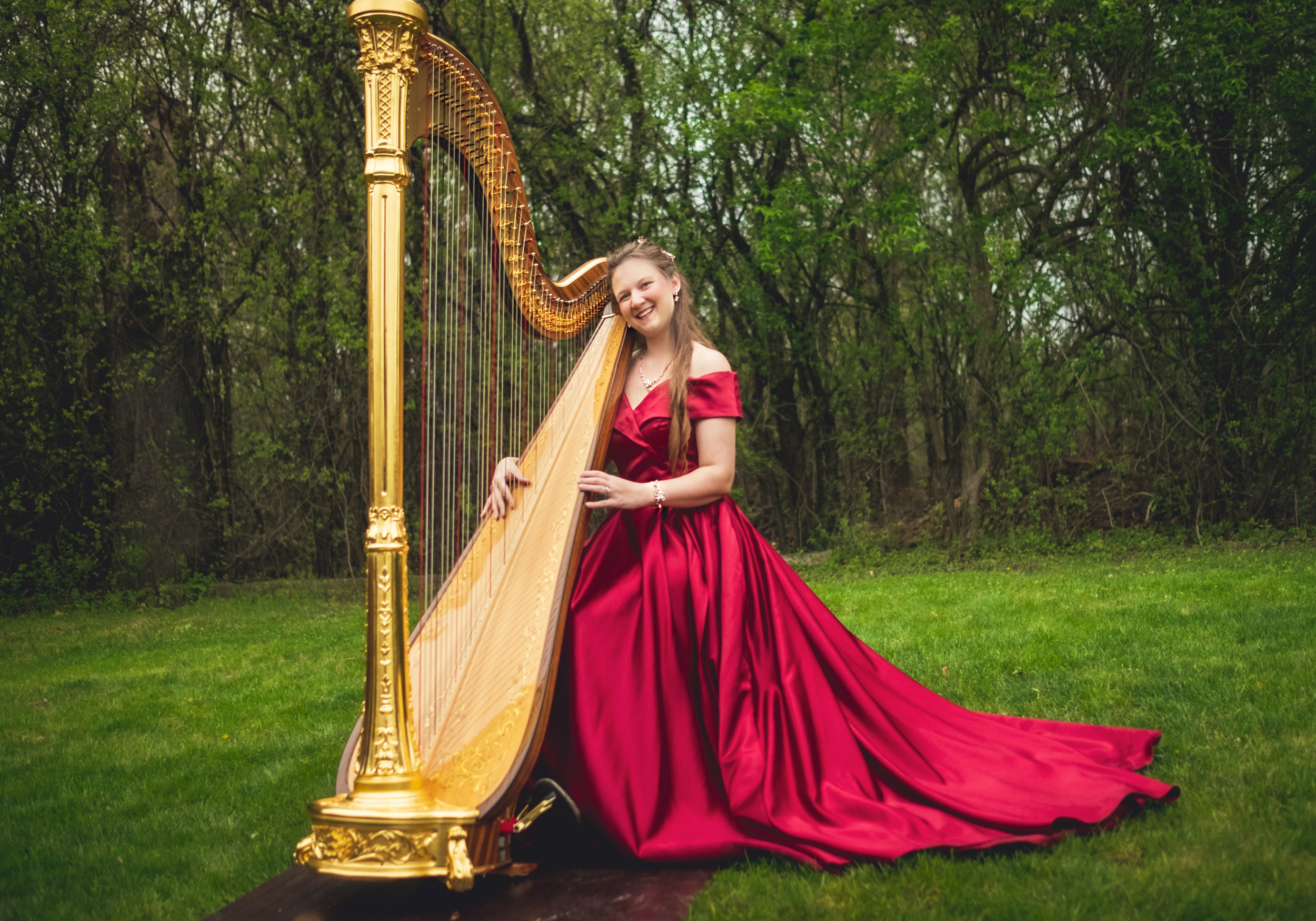 A smiling woman in a flowing red gown sits on the grass in front of a golden harp, surrounded by lush green trees.