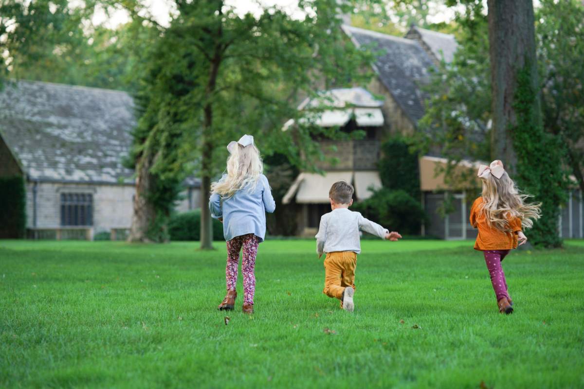 Three young children run joyfully across the green lawn at Ford House, with historic stone buildings and tall trees in the background.
