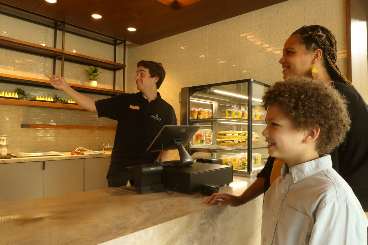 A smiling Ford House staff member behind the Speedster café counter gestures to offerings while a mother and son listen attentively. Behind them, a display case is filled with fresh grab-and-go items like salads and sandwiches.