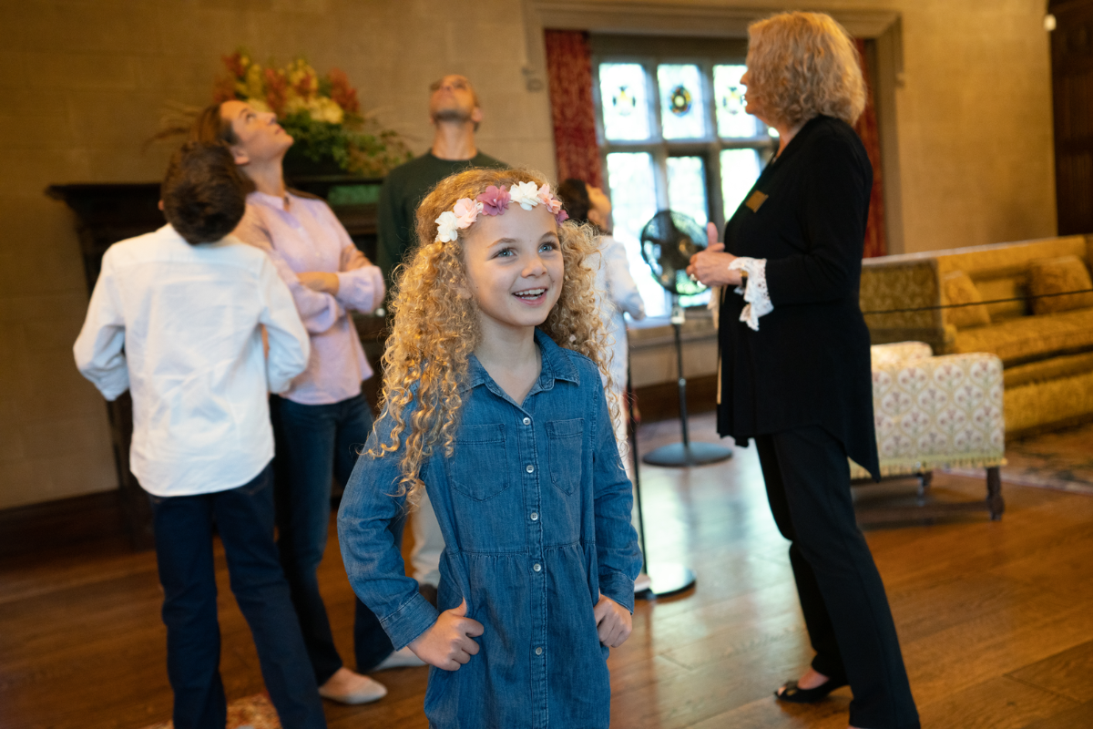 A curly-haired girl smiles brightly during a guided tour of Ford House, as adults look up at historic architectural details in the background.