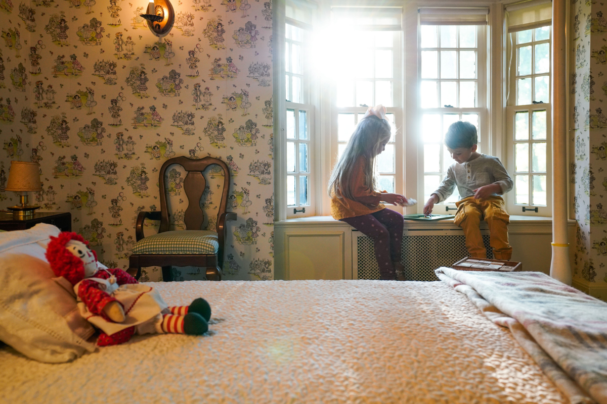 Two young children play quietly on a windowsill inside Josephine’s Playhouse at Ford House, with vintage wallpaper and soft morning light filling the space.