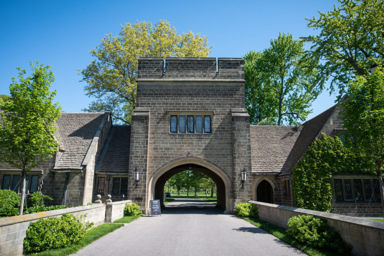 The iconic stone archway of Ford House’s main gate stands tall under a bright blue sky, framed by lush greenery and Tudor-style architecture.