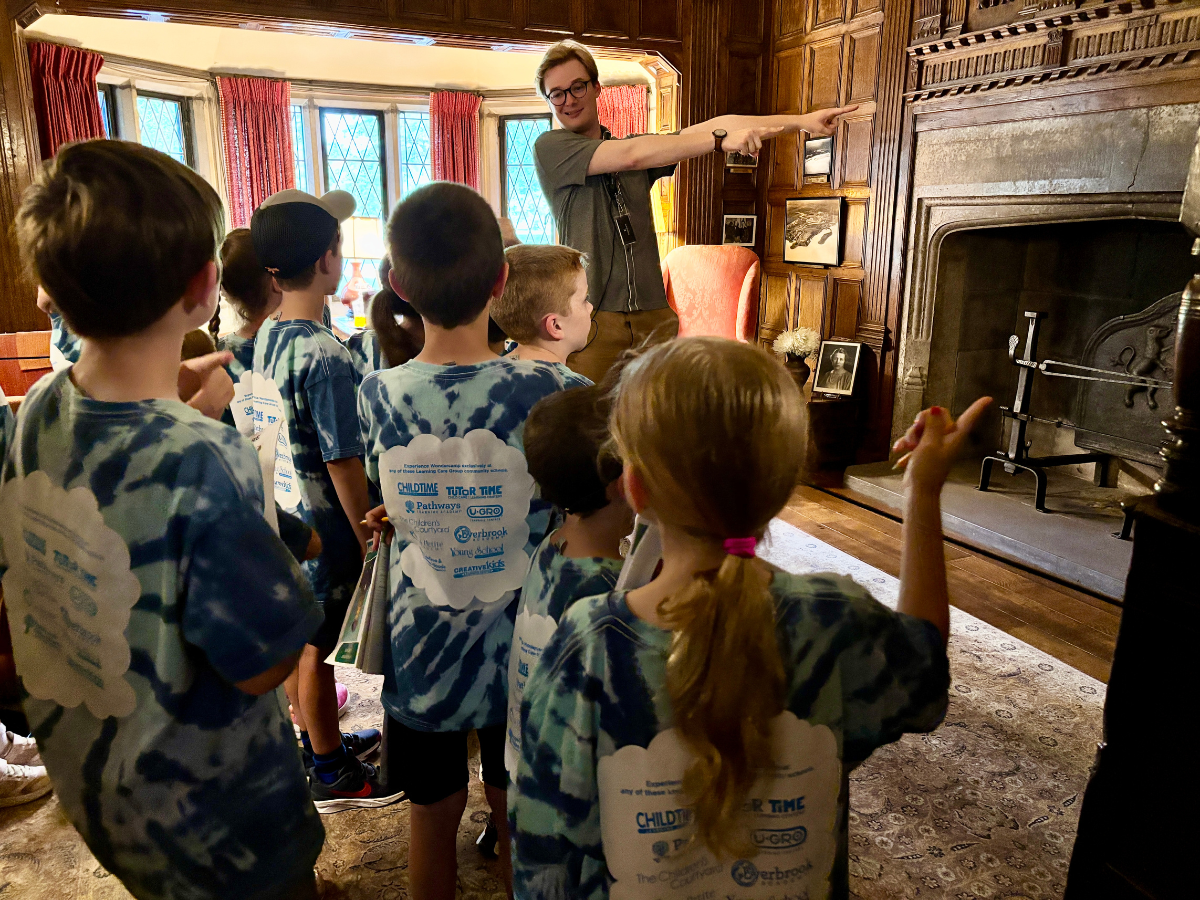 A Ford House educator leads a group of children on a school tour inside the historic Main Residence, pointing toward a detail above the fireplace as students listen attentively.