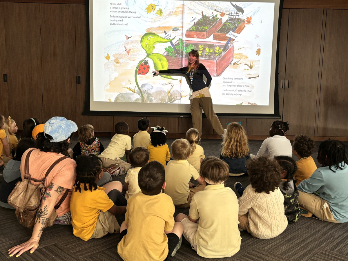 A Ford House educator reads and points to a large illustrated page projected on a screen, with children sitting cross-legged on the floor during a school program.