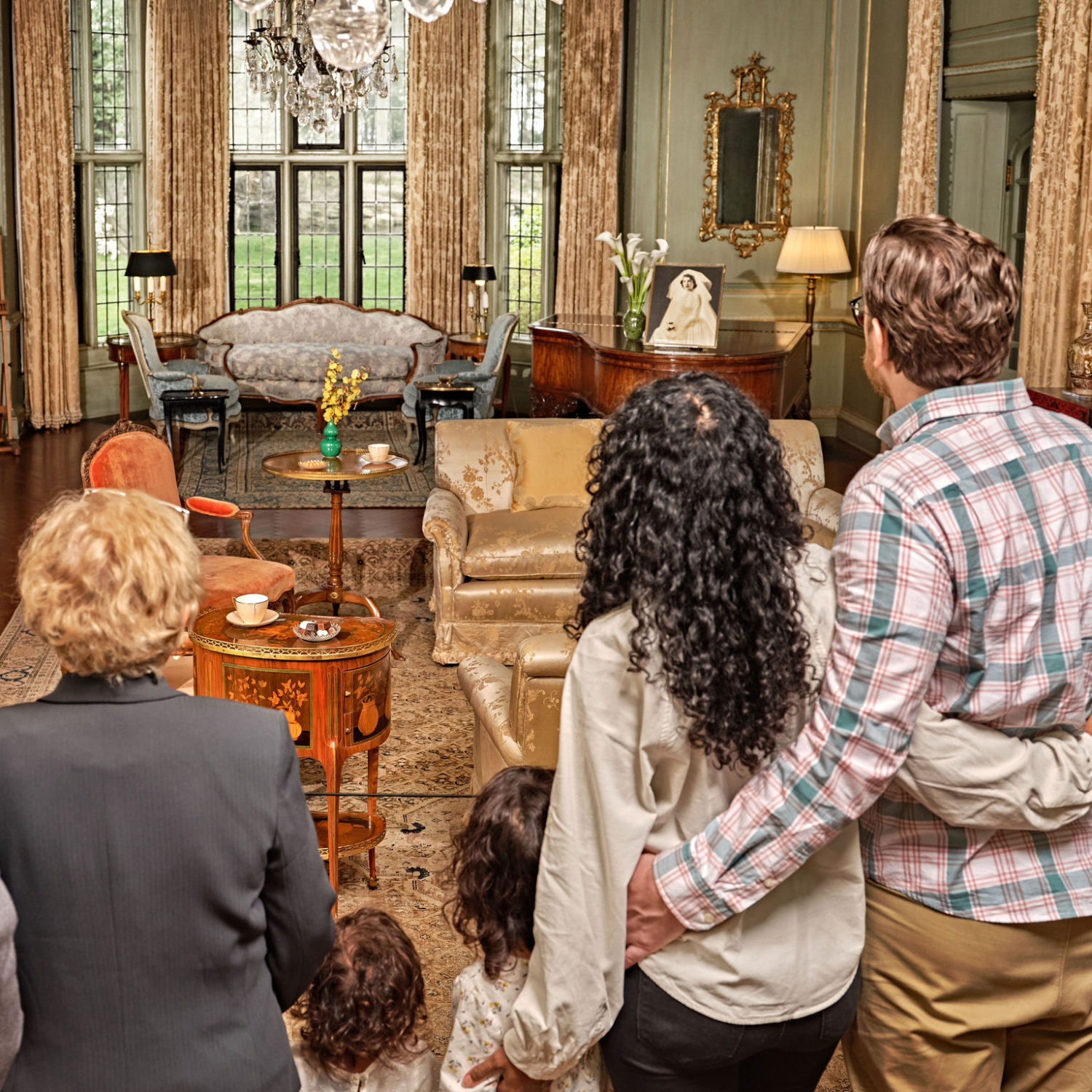 A group of visitors, including adults and children, stand together inside a grand, elegantly furnished room at Ford House, listening to a tour guide just out of frame.