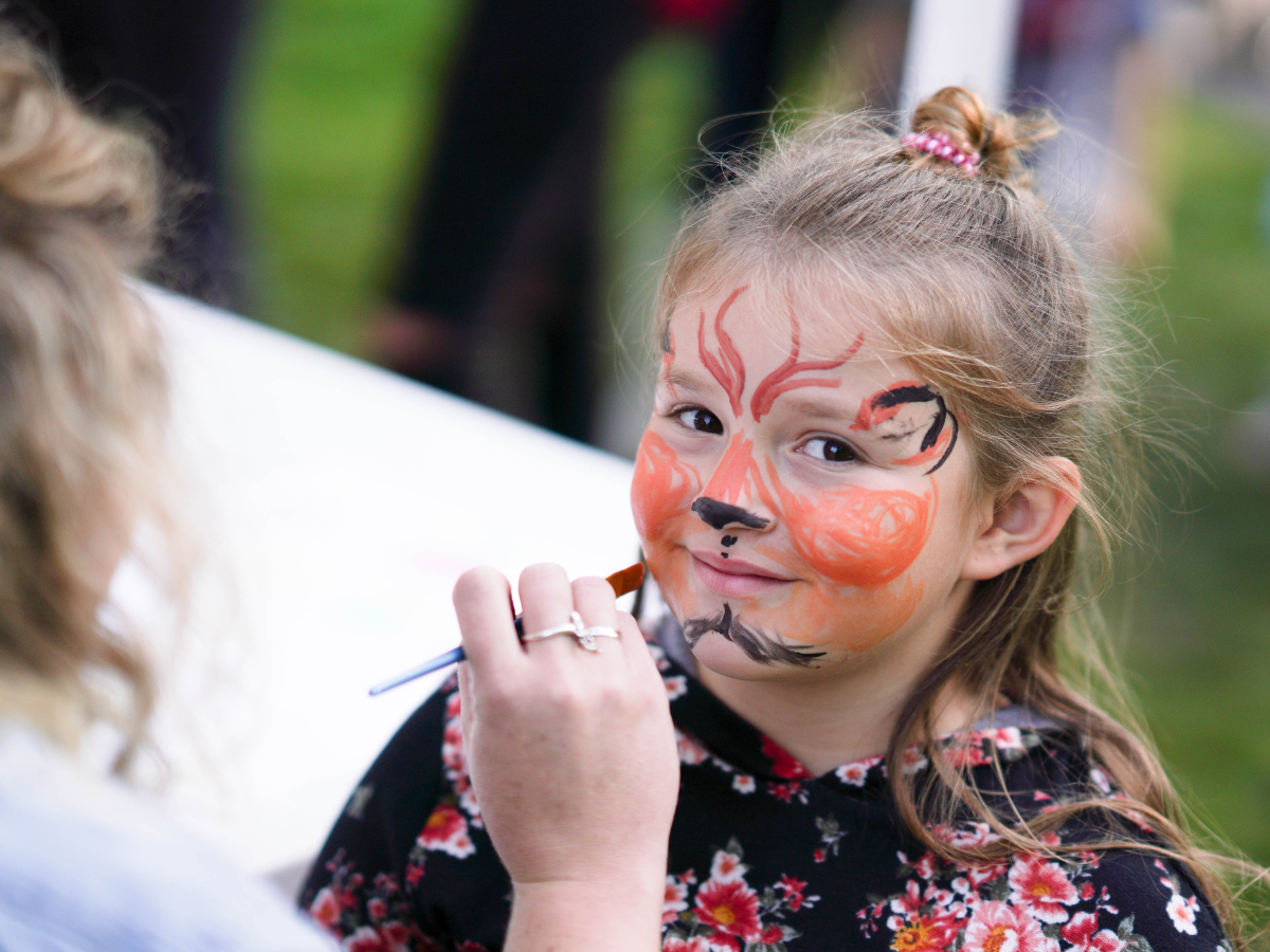 Young girl having her face painted as a tiger at Harvest Day, smiling as the artist finishes the details.