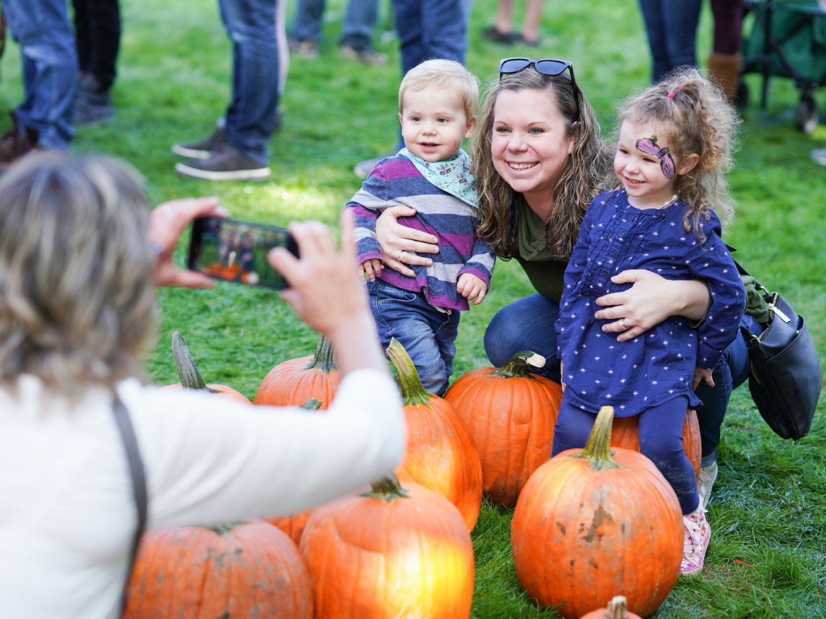 Mother posing with two children among a display of pumpkins at Harvest Day, while another adult takes their photo.