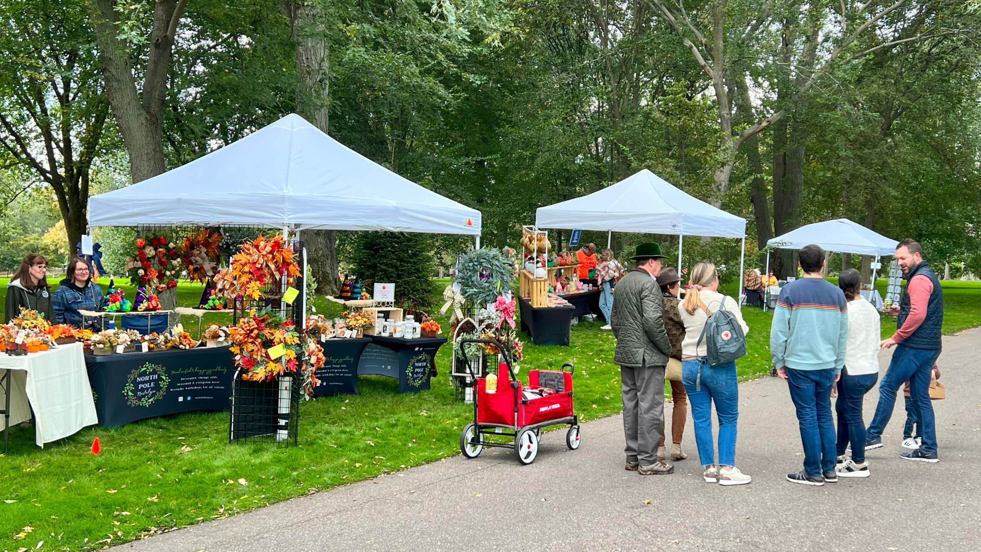 Shoppers explore fall-themed vendor booths under white tents, showcasing handmade wreaths, autumn decorations, and artisan goods along a tree-lined path.