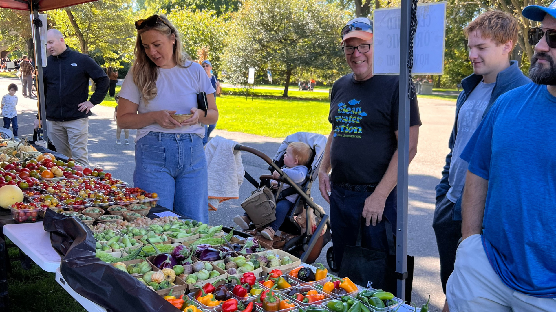 A woman shops for fresh produce at an outdoor vendor table filled with colorful tomatoes, peppers, and greens as a baby sits in a stroller nearby and other guests look on.