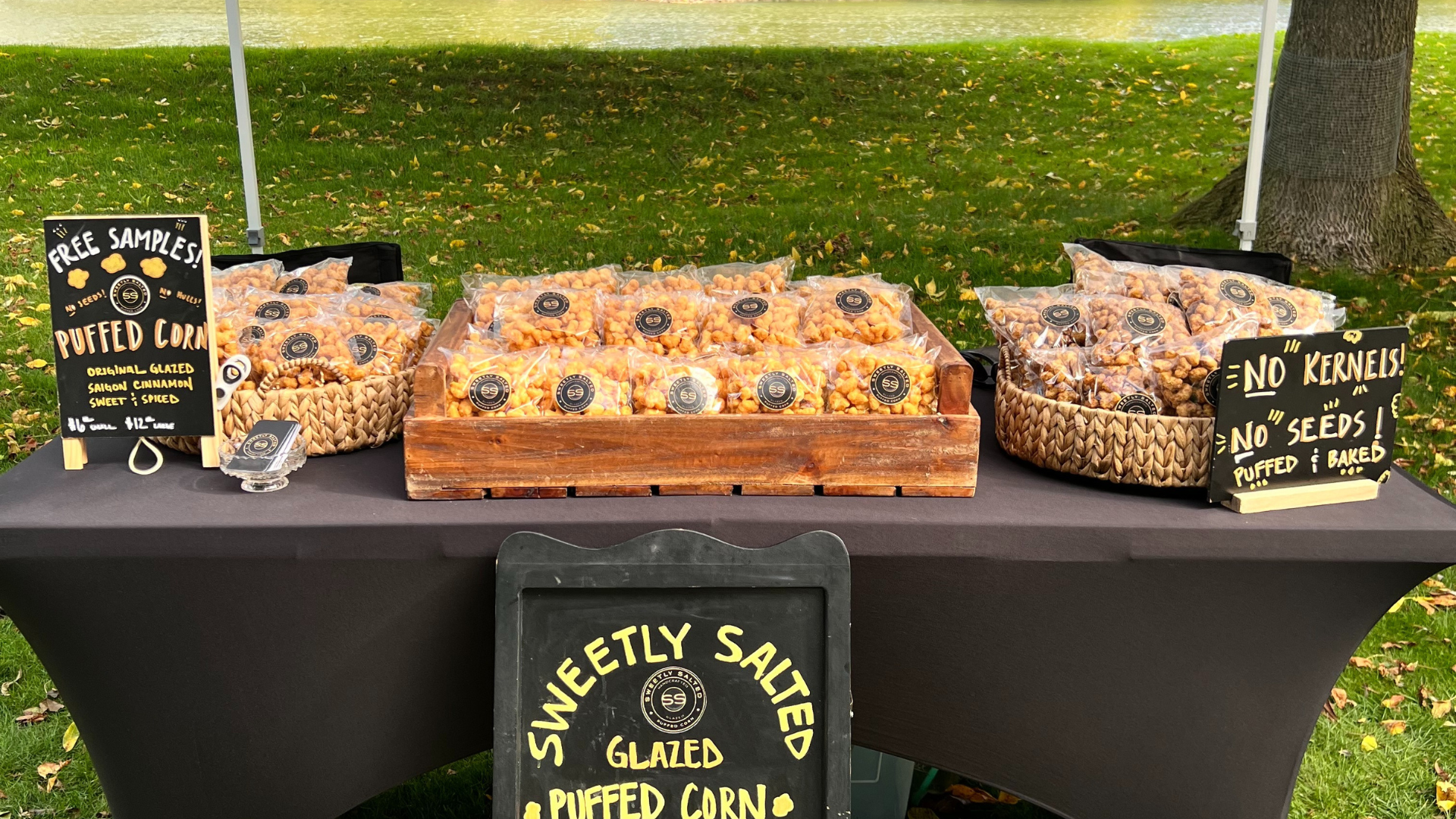 Bags of puffed corn snacks are neatly arranged on a vendor table with signs offering free samples and highlighting flavors like sweet, spiced, and cinnamon.