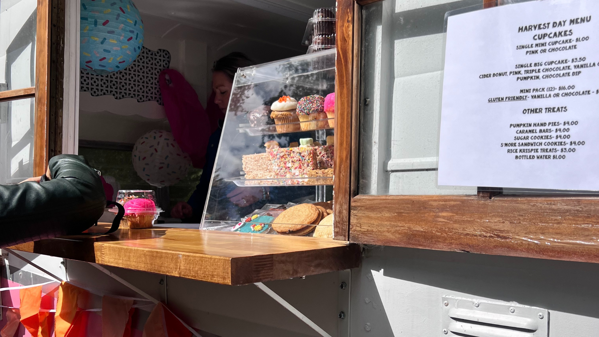 A display case filled with cupcakes, cookies, and colorful sweets is set up in a food truck window at Ford House’s Harvest Day, with a printed menu beside it.