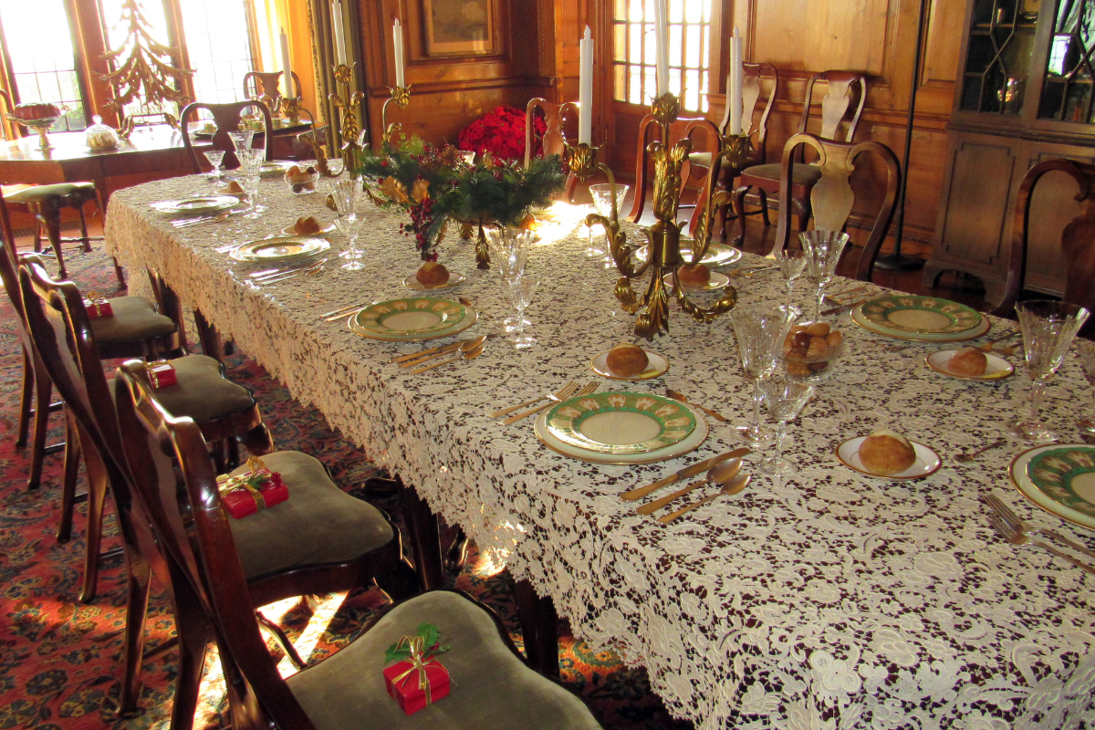 Elegant formal dining room table at Ford House set with lace tablecloth, gold candelabras, fine china, crystal glasses, and small wrapped gifts at each place setting for Home for the Holidays.