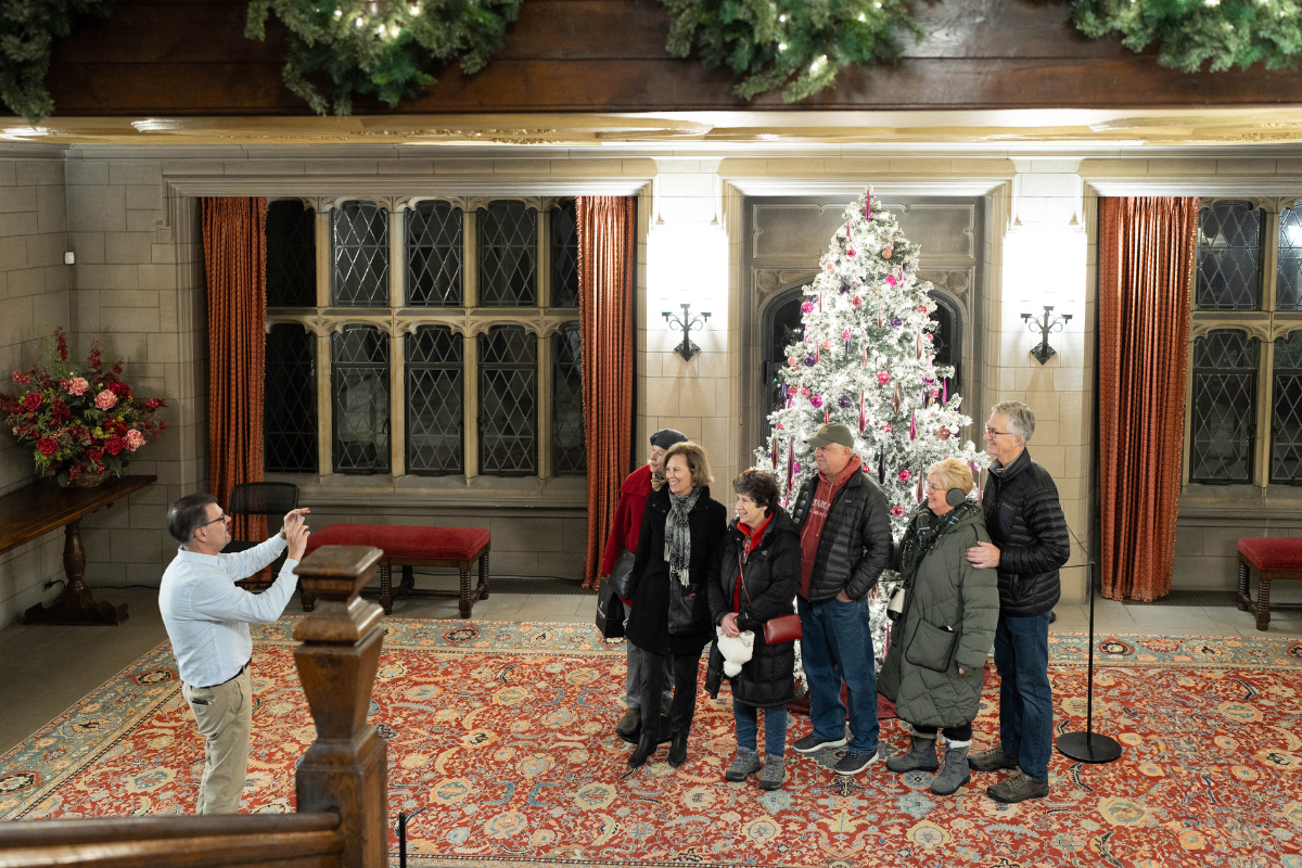 A group of six guests pose in front of a white Christmas tree with pink and silver ornaments inside the Main Residence at Ford House, while another guest takes their photo.