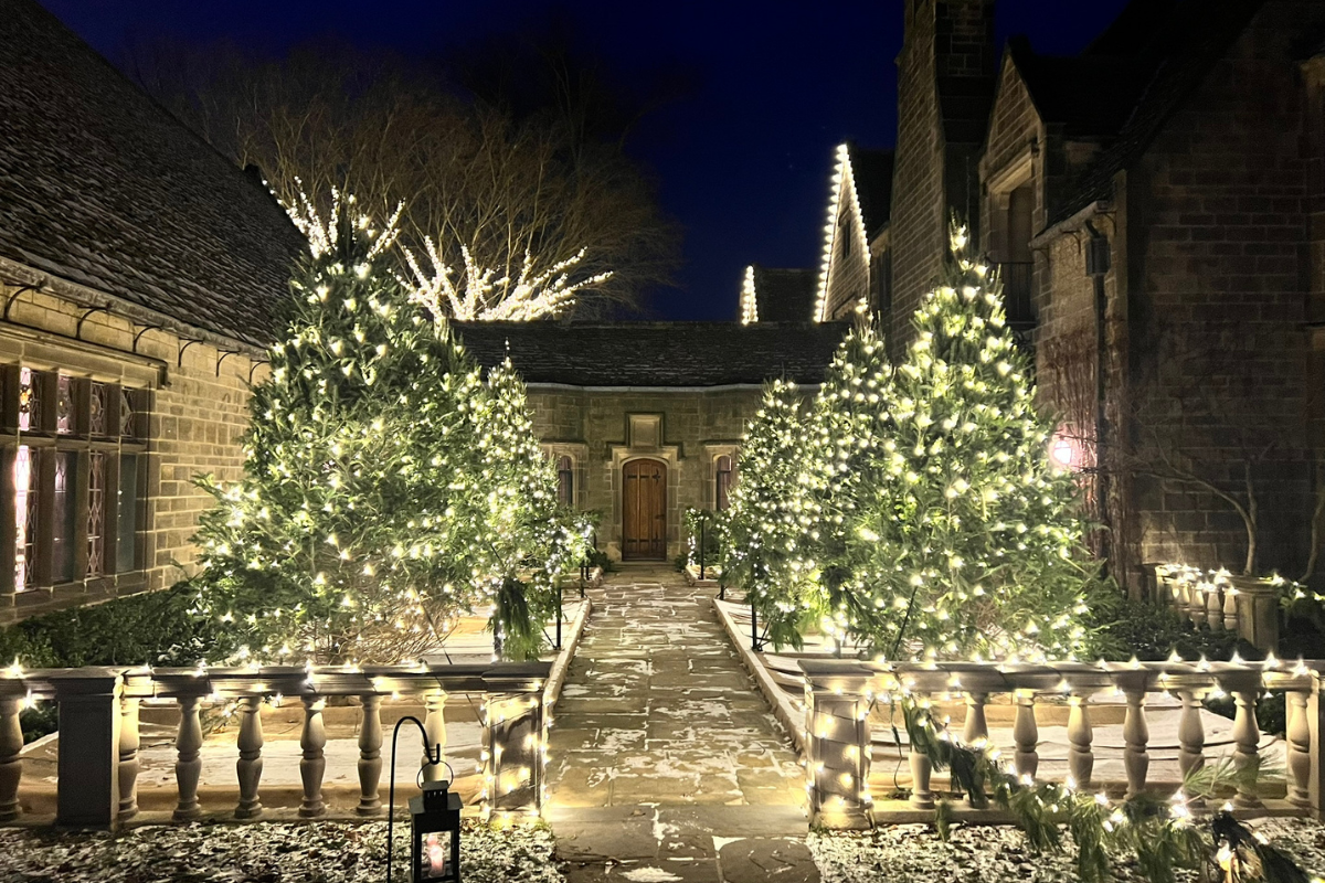 A stone courtyard lined with potted evergreen trees wrapped in white lights, leading to a wooden door at Ford House during Home for the Holidays.