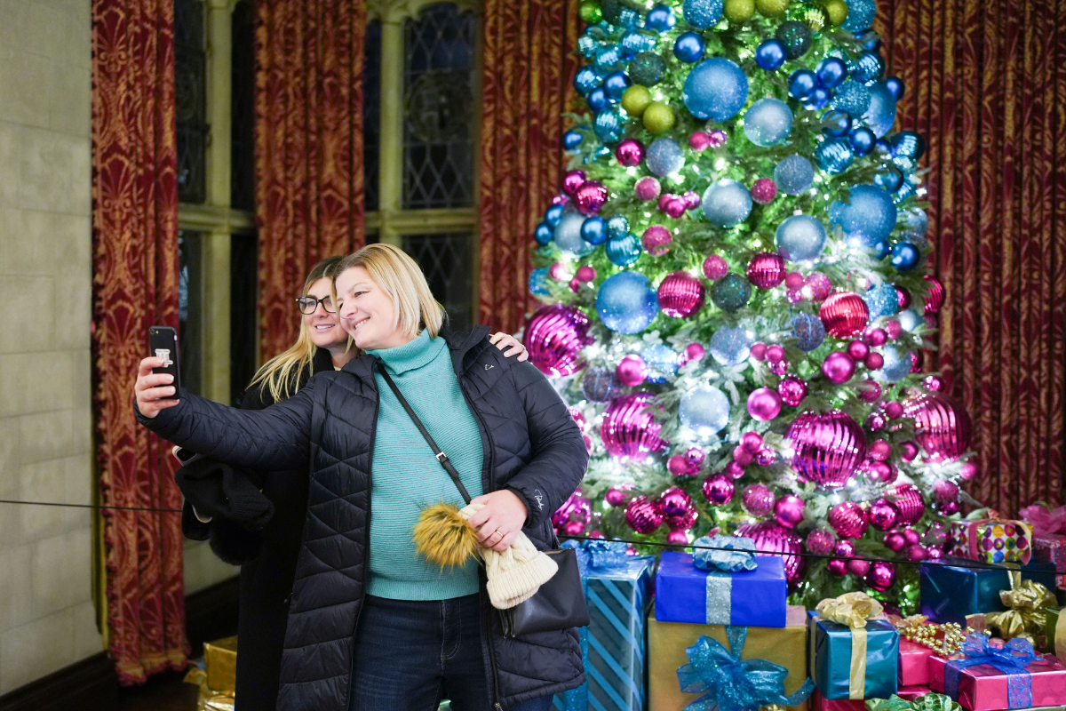 Two women smile for a selfie in front of a tall Christmas tree decorated with blue, pink, and green ornaments inside the Main Residence at Ford House.