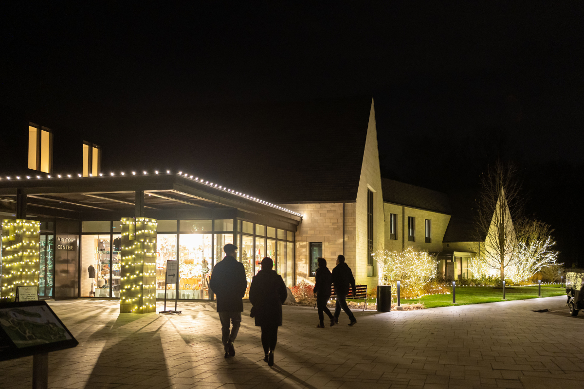 Guests walk toward the Ford House Visitor Center at night, decorated with soft white holiday lights.