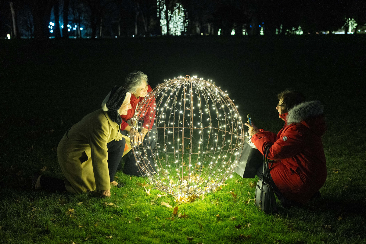 Three guests gather around a large spherical light sculpture on the lawn during Home for the Holidays at Ford House.