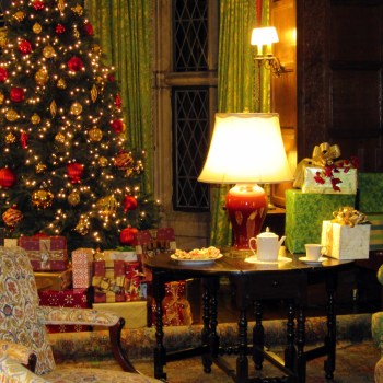 A warmly lit room at Ford House decorated for Home for the Holidays at Ford House, featuring a Christmas tree, wrapped presents, and vintage furniture.