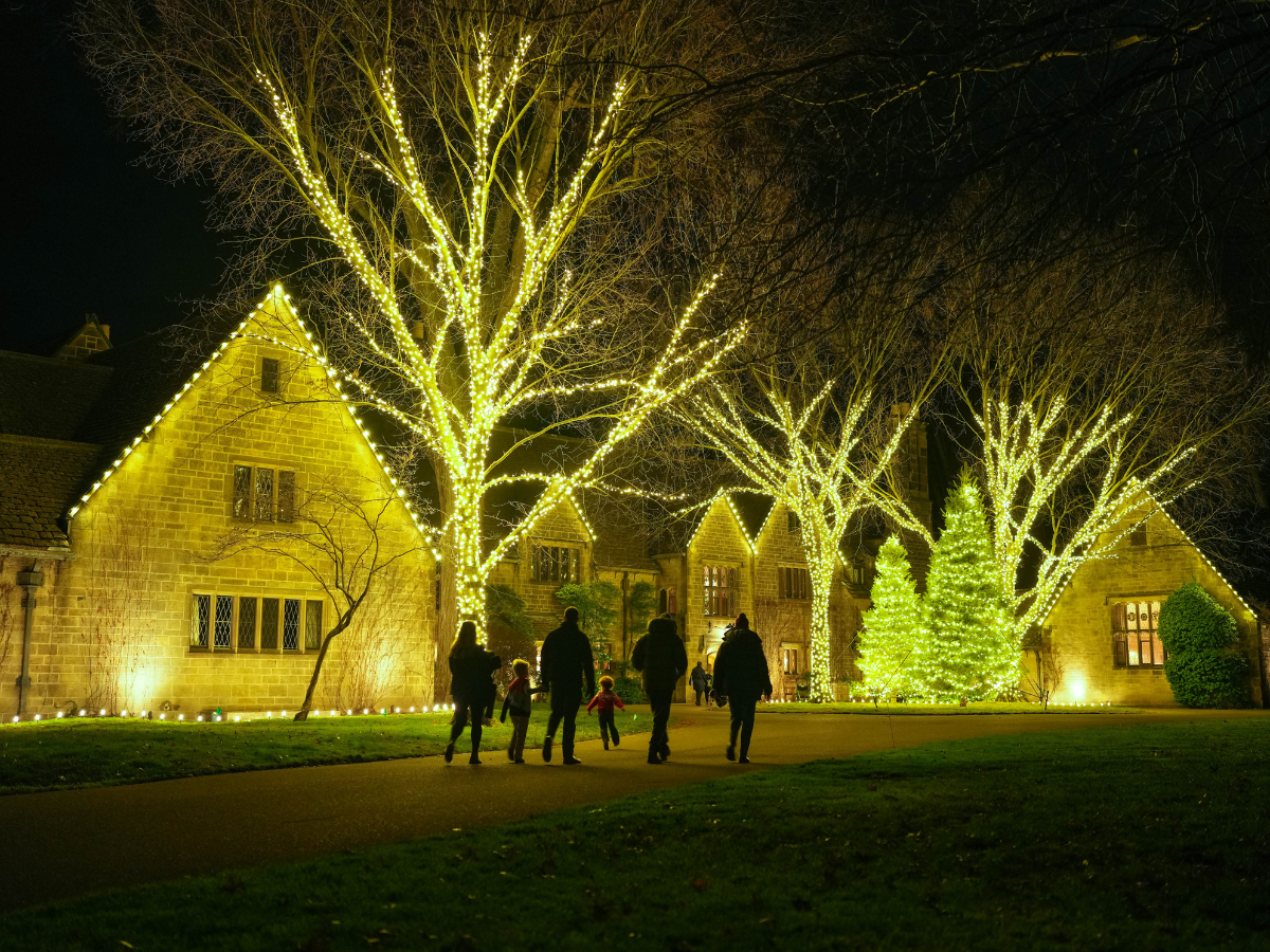 Families walk toward the warmly lit Main Residence at Home for the Holidays at Ford House, surrounded by trees and evergreens wrapped in golden holiday lights.