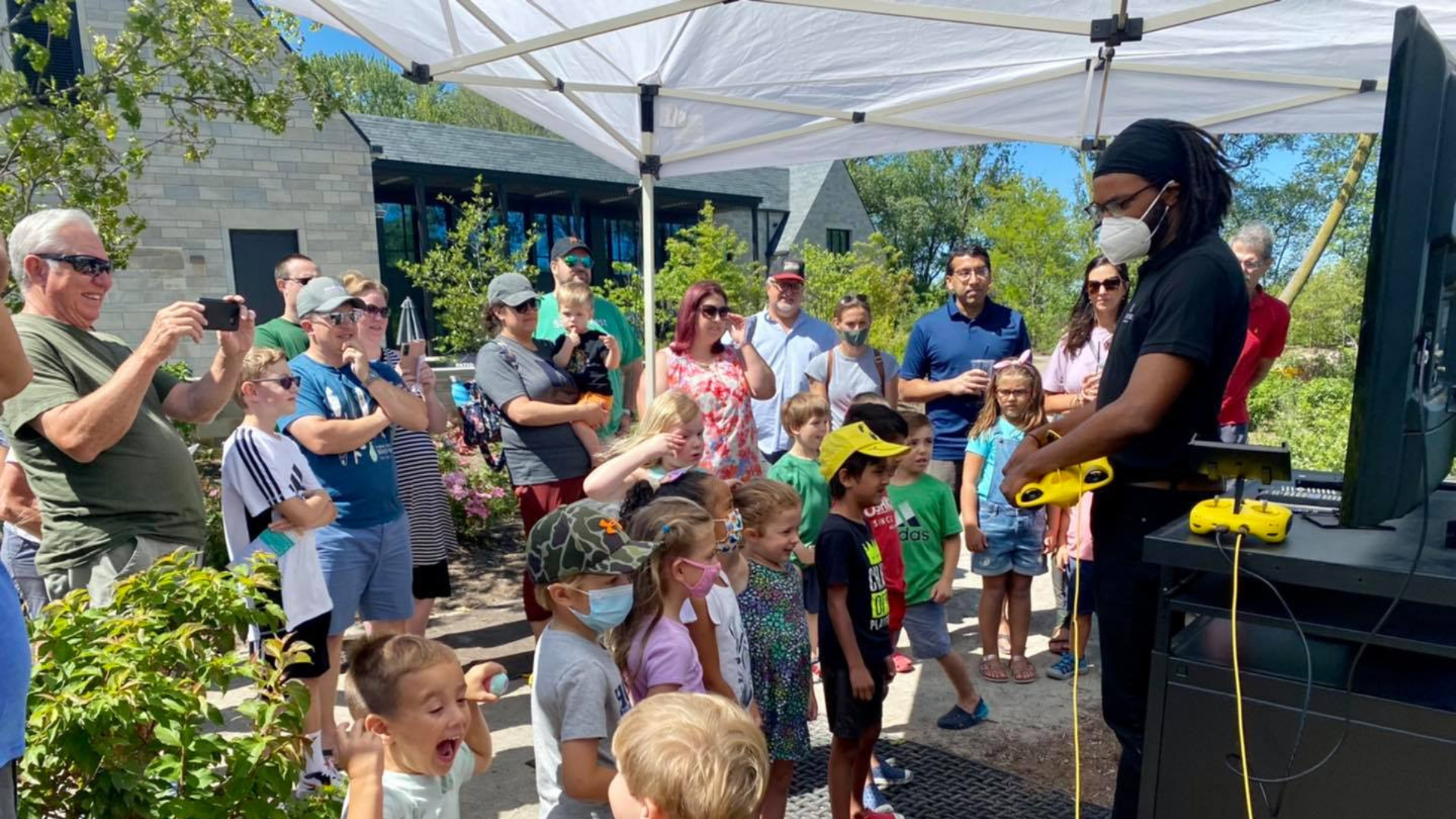 A crowd of families and children gathered outdoors under a white canopy, watching a demonstrator operate underwater drone equipment.