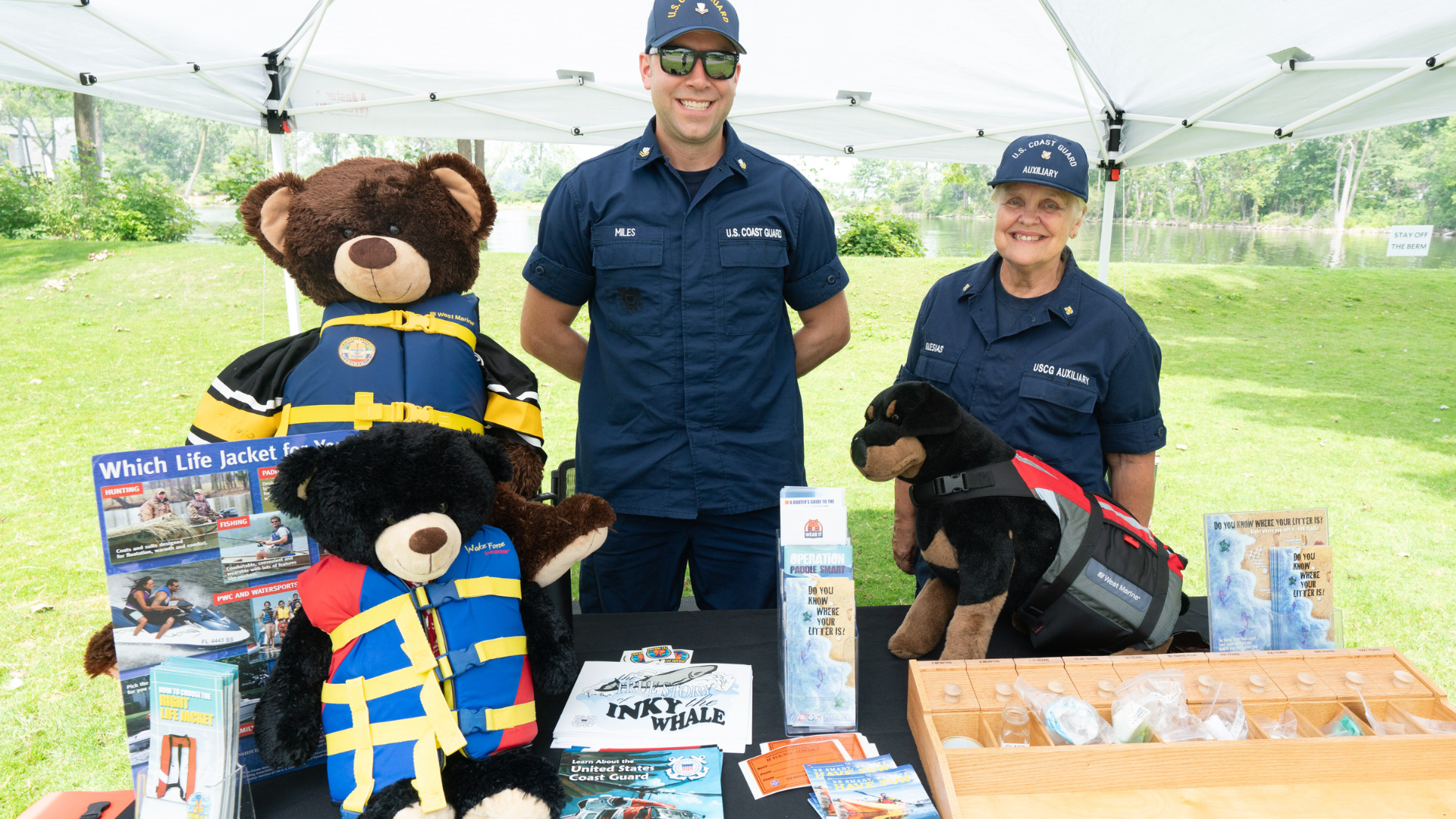 Two U.S. Coast Guard Auxiliary members stand behind a booth with stuffed animals in life jackets and water safety information.