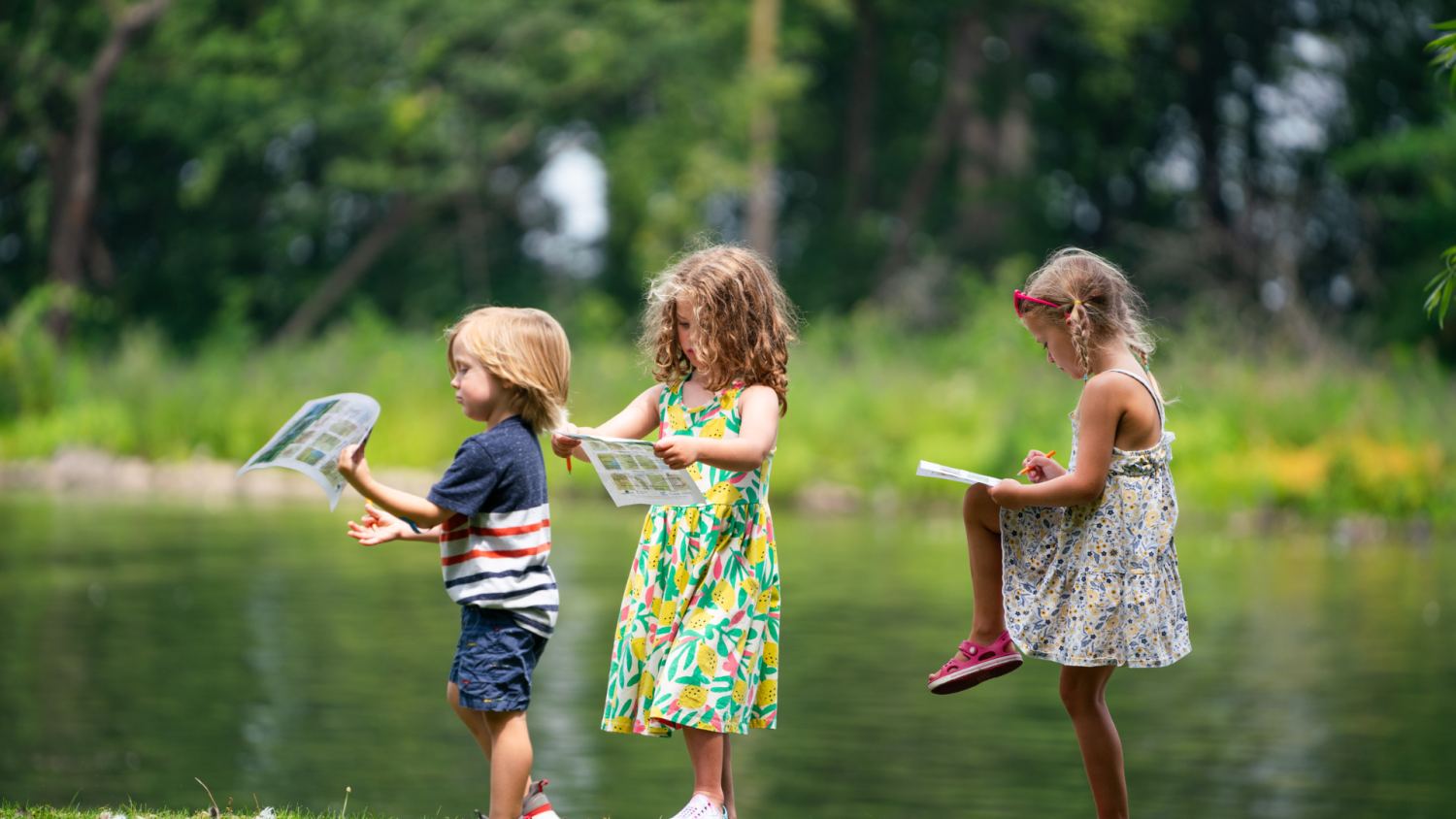 Three young children standing near a pond, holding activity sheets and pencils as they explore nature.