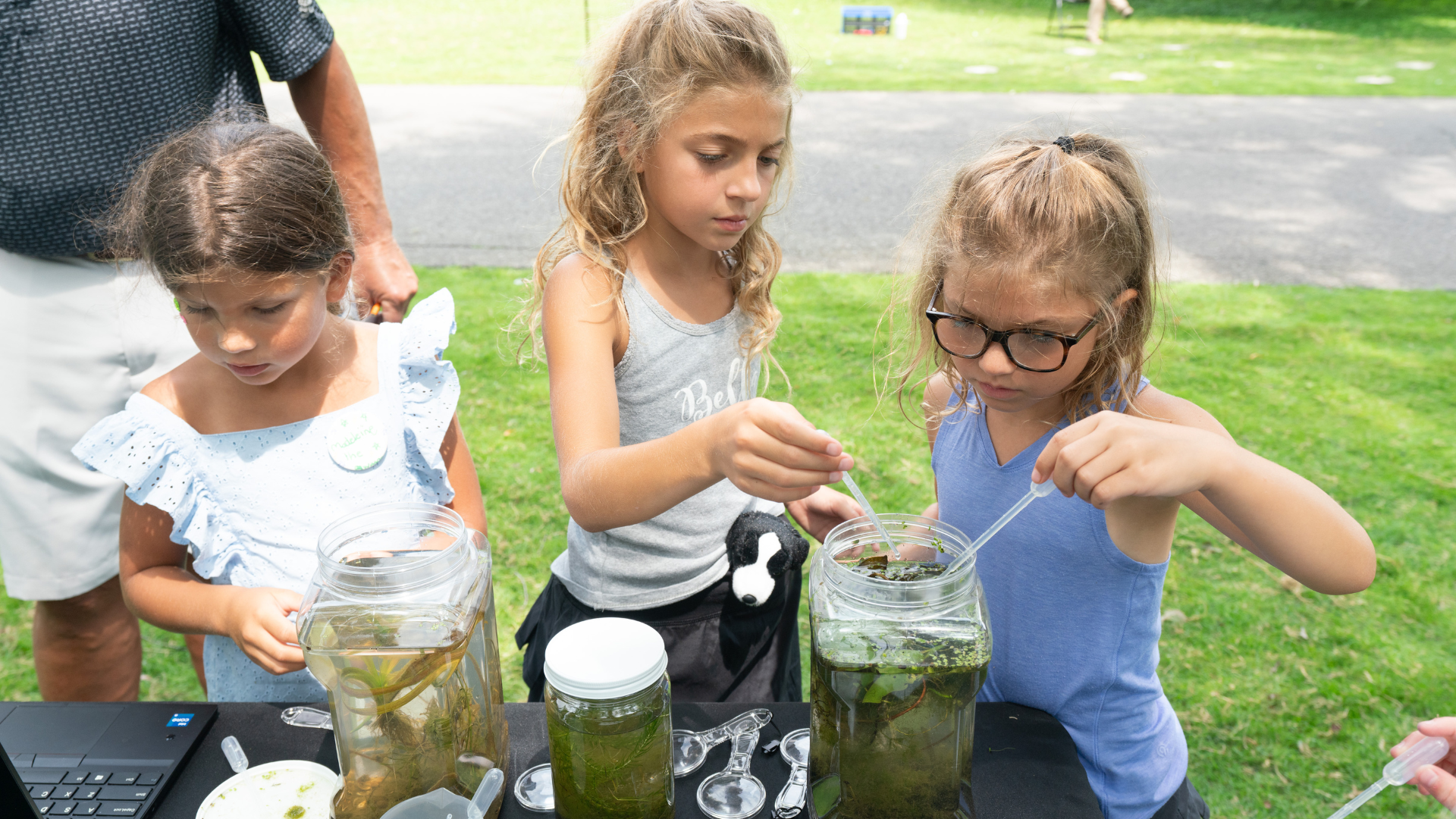 Three girls closely examining jars of pond water and plants using droppers and tools at an outdoor science activity table.