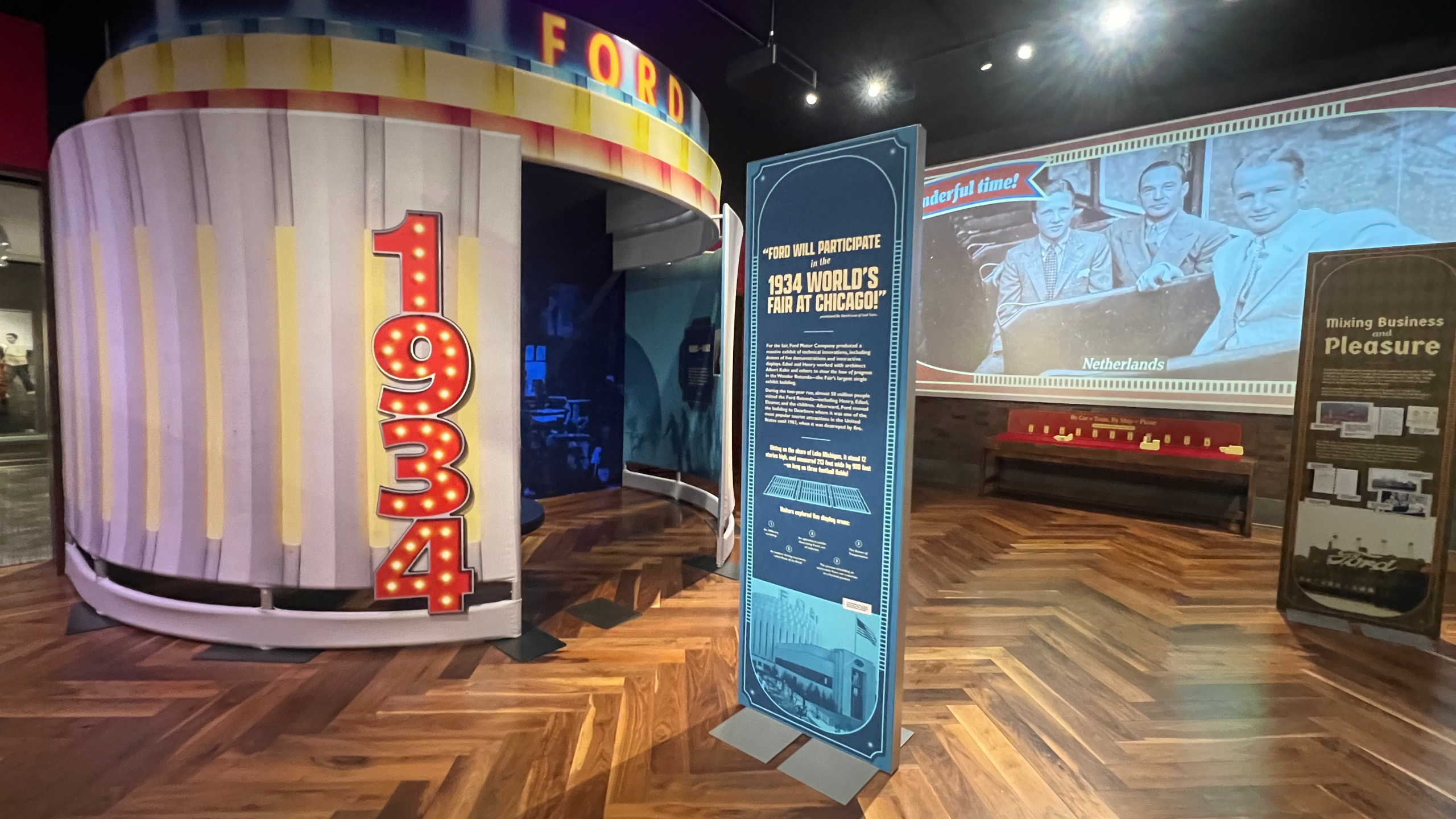 A 1934 World’s Fair-themed section of the Ford family travel exhibit, featuring a tall blue panel describing the Ford Motor Company’s participation in the Chicago fair. The background includes a curved art deco-style structure with colorful stripes and the word “FORD,” as well as projected photos of Edsel Ford and companions in the Netherlands.