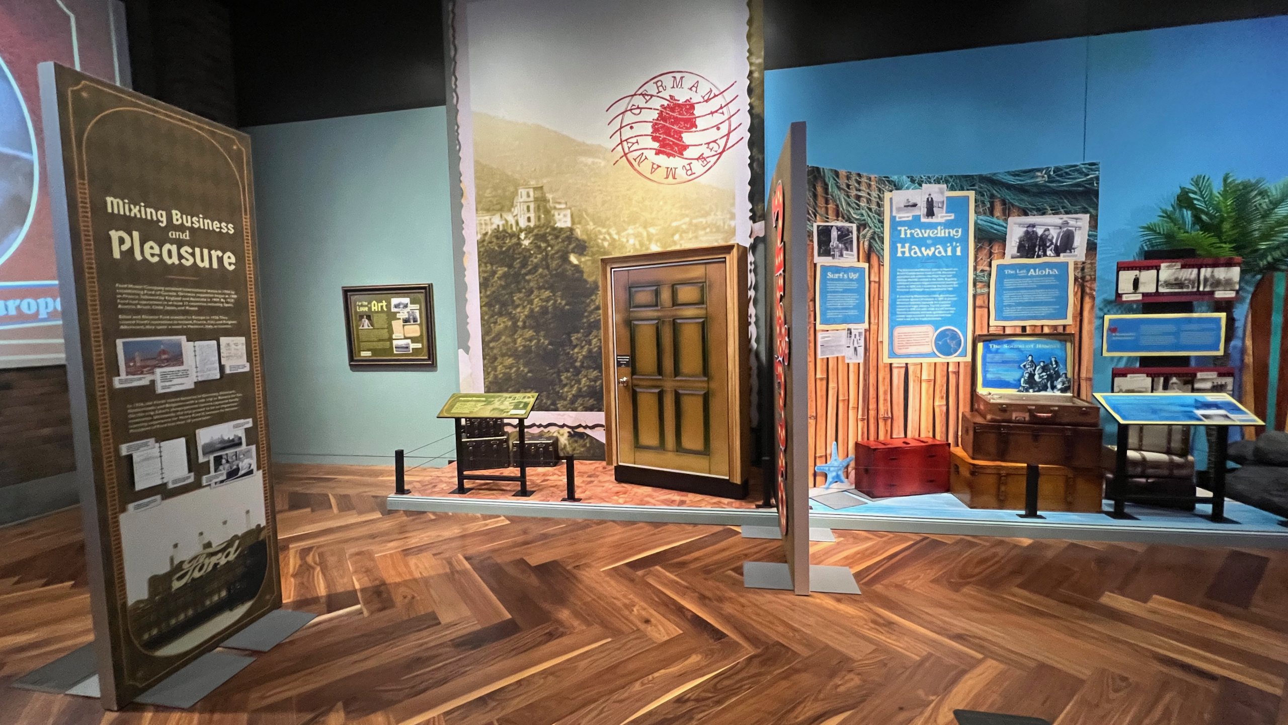 The “Mixing Business and Pleasure” display in the Ford family travel exhibit, flanked by scenes from Germany and Hawai’i. The exhibit features a backdrop with a large vintage photo of European landscapes and a door suggesting immersive entry into a new scene. Panels explain the blending of professional and leisure travel.