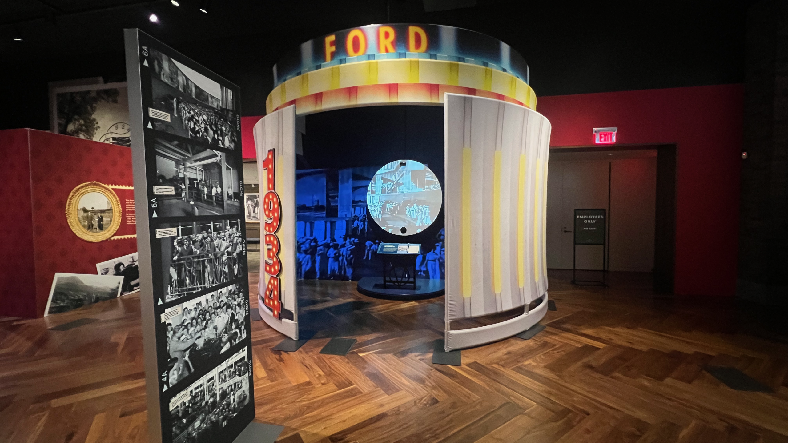 A circular screen projects black-and-white footage of Edsel Ford and others exiting a car at the 1934 World’s Fair, surrounded by blue-tinted historic images of vintage automobiles and spectators. A small display stand with descriptive text sits in front of the screen.