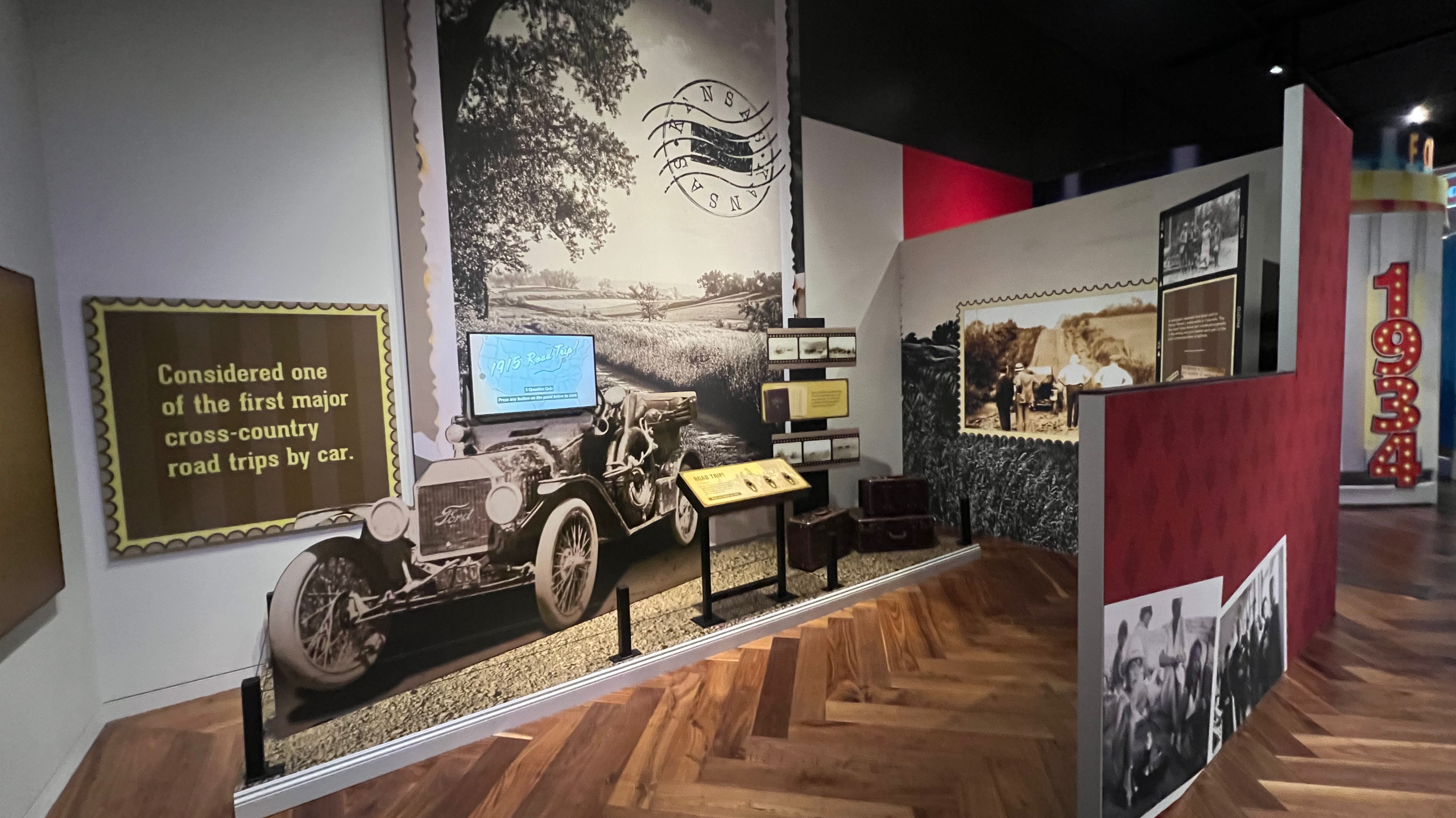 A transitional view from the 1915 road trip section to the Hawai’i display, with panels showing historic photographs, quotes from travelers, and a bold red sign labeled “Better Cars, Better Drivers” standing beneath the “Wish You Were Here!” banner.