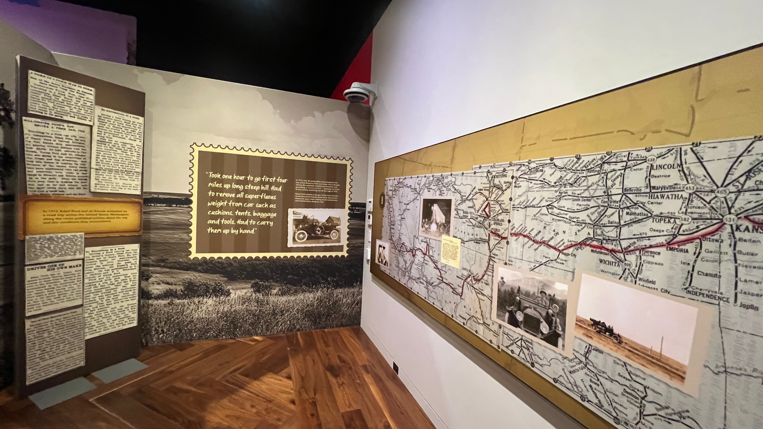 Wall exhibit at Ford House displaying the Ford family’s 1915 cross-country road trip, featuring vintage newspaper clippings, a large quote about climbing steep hills, a sepia-toned photo of a Model T, and a large map with a red travel route marked across the Midwest. Additional photos of early automobiles are pinned to the map.