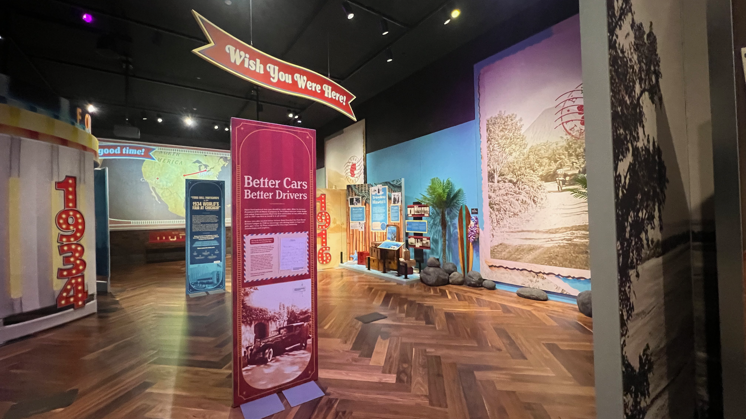 Wide view of the Wish You Were Here! exhibit showing the “Better Cars Better Drivers” panel in the center, flanked by a curved “1934” World’s Fair display on the left and a tropical-themed 1916 Hawai‘i section on the right. A red arrow-shaped banner with the exhibit title hangs above. Surfboards, luggage, palm trees, and vintage travel graphics create an immersive environment.