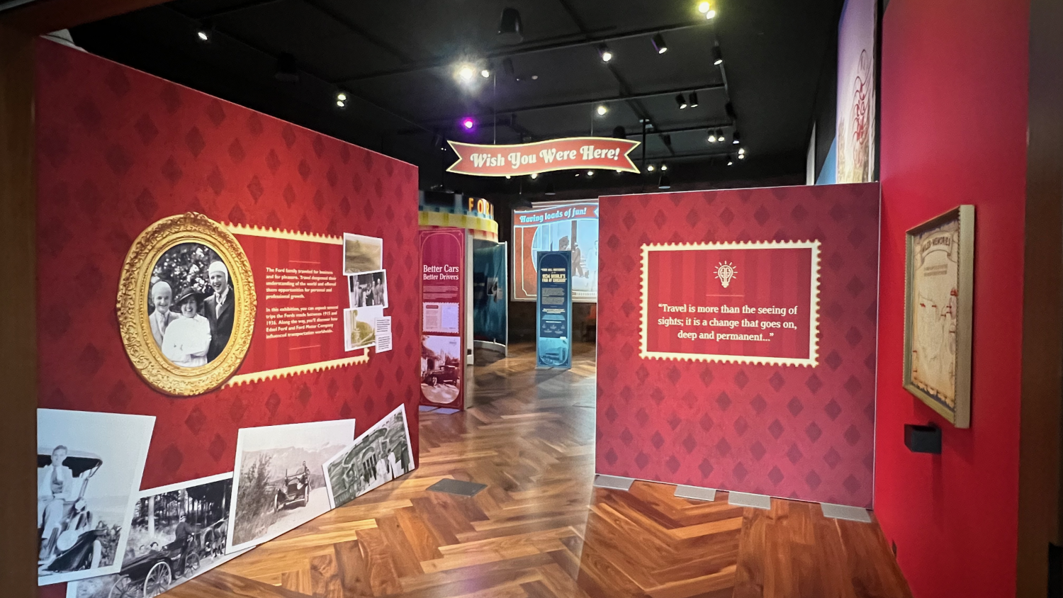 Entrance to the “Wish You Were Here! Unpacking Ford Family Travel” exhibit at Ford House, featuring a red wall with vintage photos of the Ford family, a gilded portrait, and text introducing the family’s historic travels between 1915 and 1934. A decorative banner reading “Wish You Were Here!” hangs above the exhibit entryway.