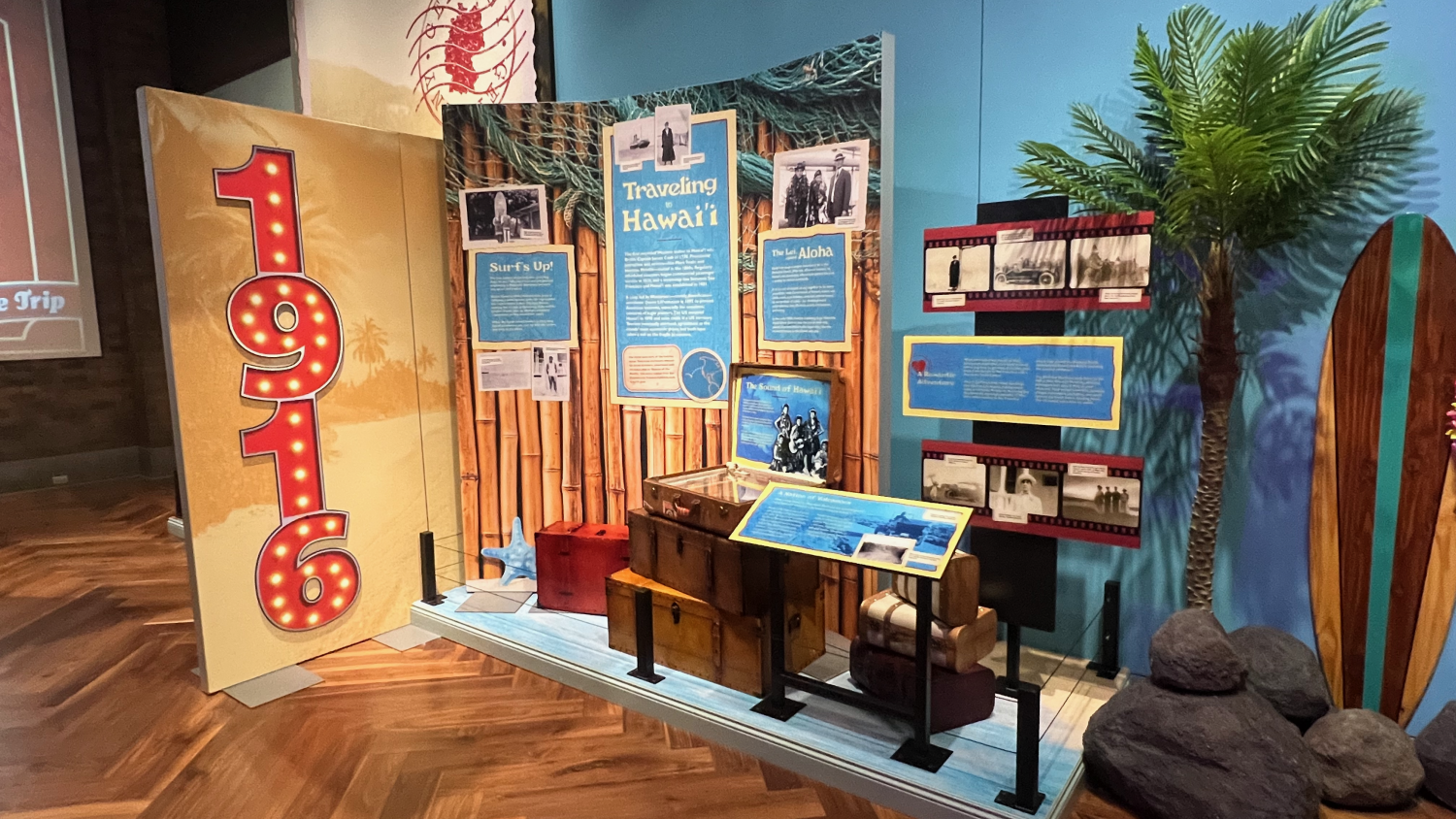 The “Traveling to Hawai’i” section of the Ford family travel exhibit, decorated with bamboo textures, tropical visuals, and luggage props. The display includes vintage photographs, informational panels on surfing, leis, and Hawaiian fashion, with a surfboard and palm trees adding to the immersive island atmosphere.