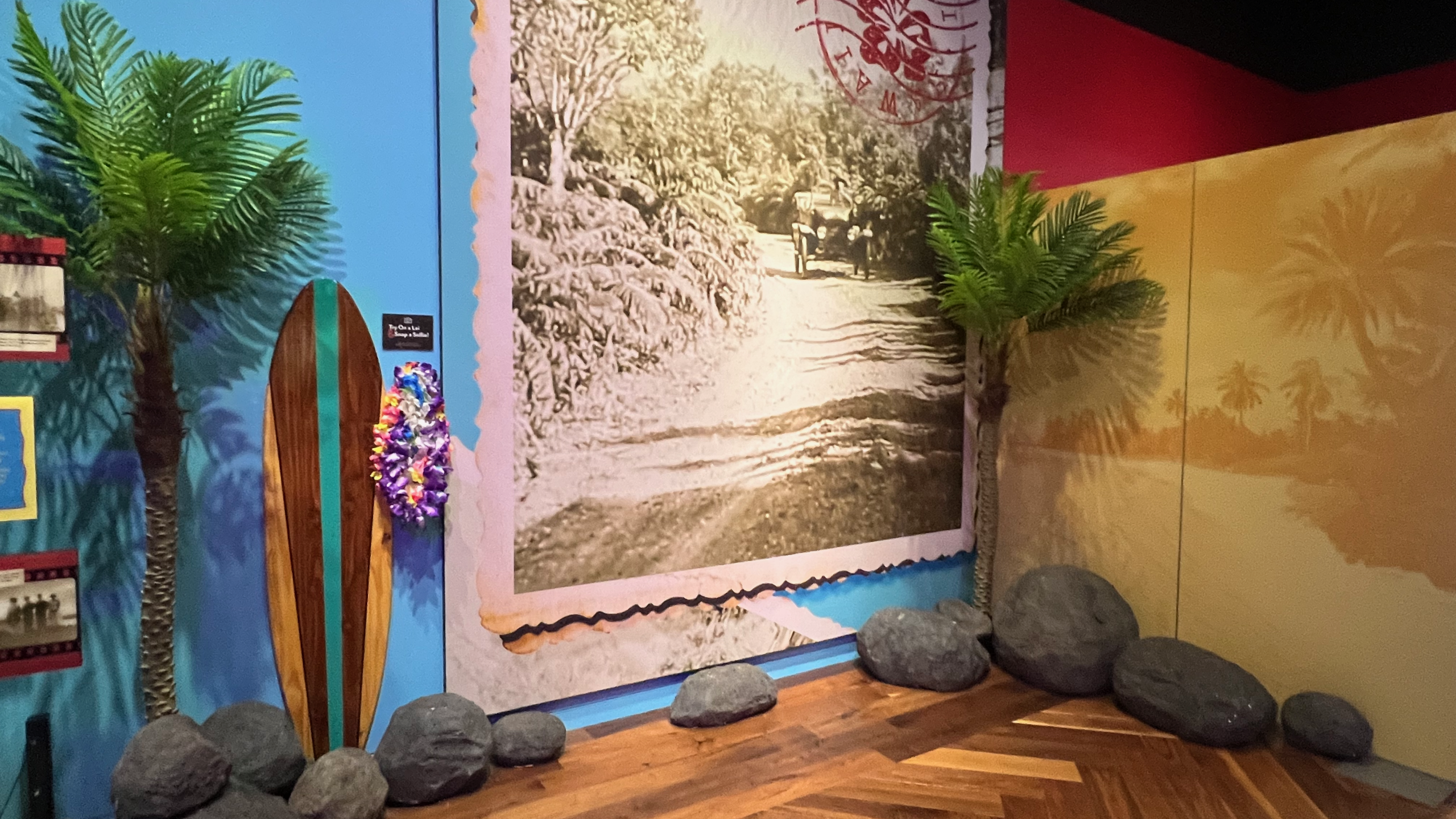 Corner of the Hawai’i-themed section of the Ford family exhibit, featuring a sepia-toned photograph of a carriage on a Hawaiian road. Artificial palm trees and faux lava rocks enhance the tropical setting, along with a wooden surfboard adorned with colorful leis.